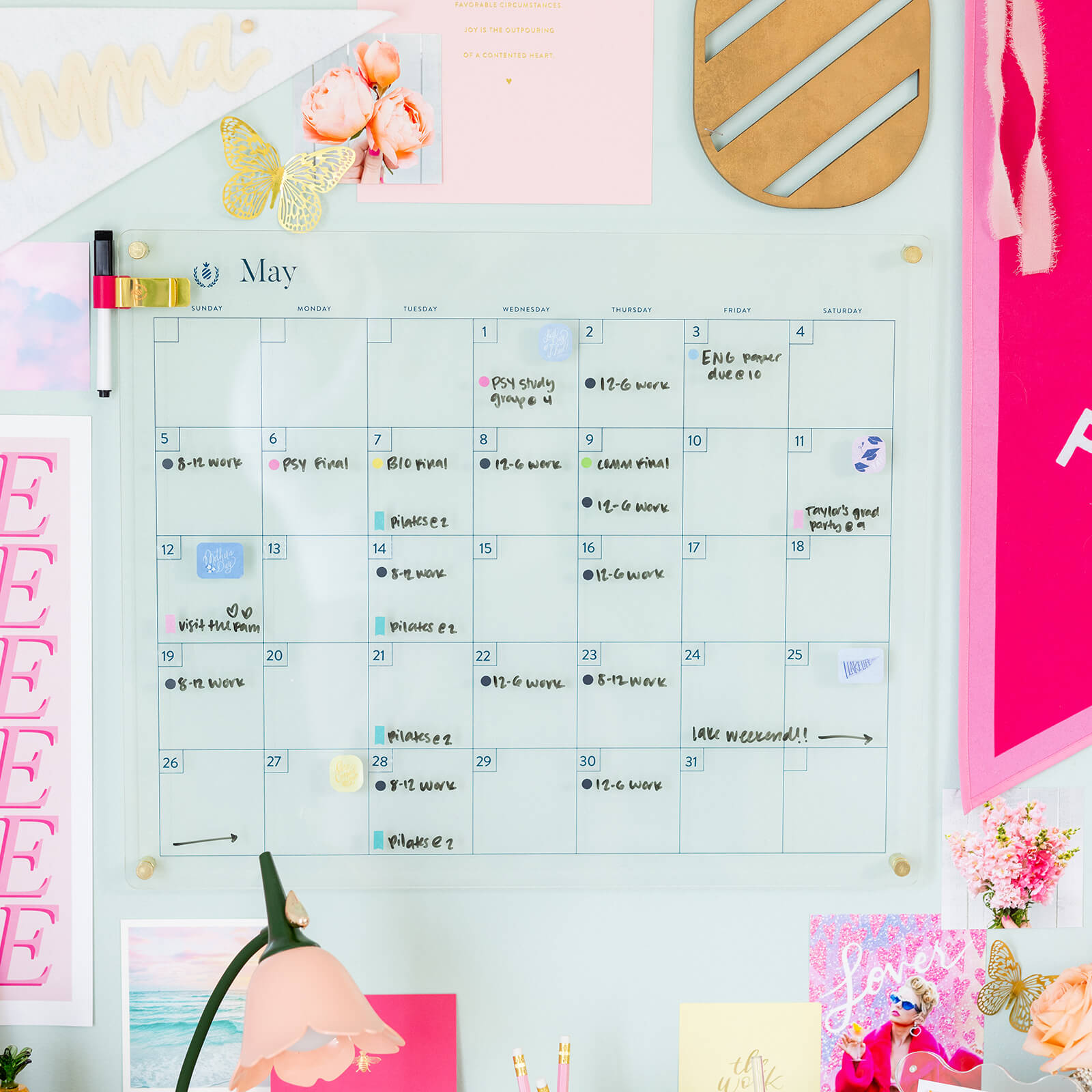 The Simplified Monthly Acrylic Wall Calendar is mounted on a wall adorned with pink notes, photos, and gold accents. Ideal for organizing events and reminders, it features handwritten plans and a marker clipped to the board.