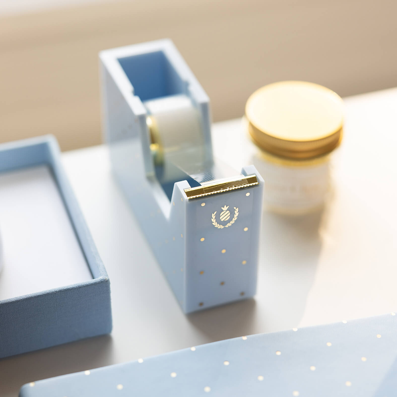The Simplified Tape Dispenser - Carolina Dot in blue with a gold foil emblem sits on a white desk next to a matching box and gold-lidded jar, softly lit for an elegant look.