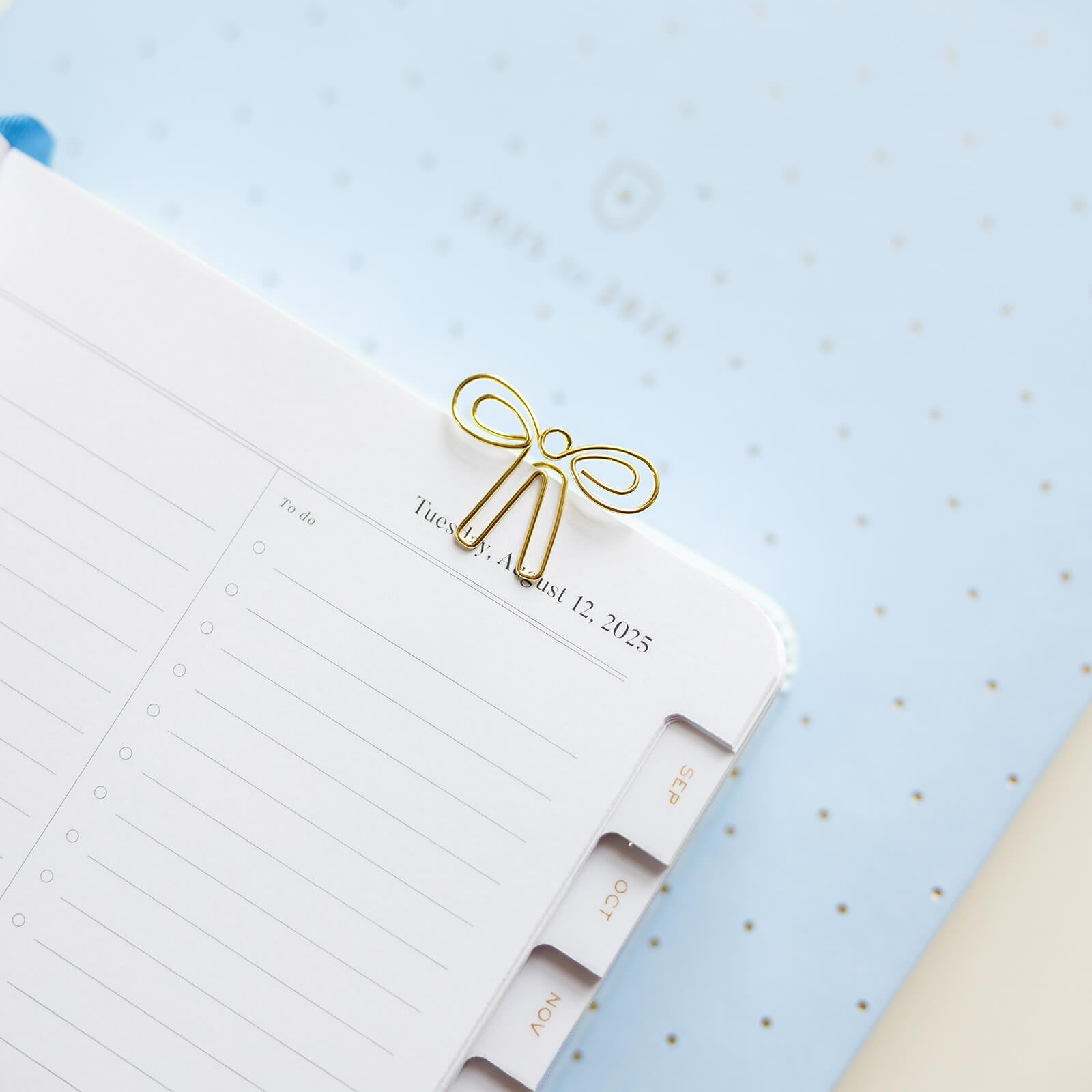 A Simplified Paper Clips - Bows securely holds a planner corner open, showing lined pages and tabbed dividers atop a pale blue polka-dotted surface.