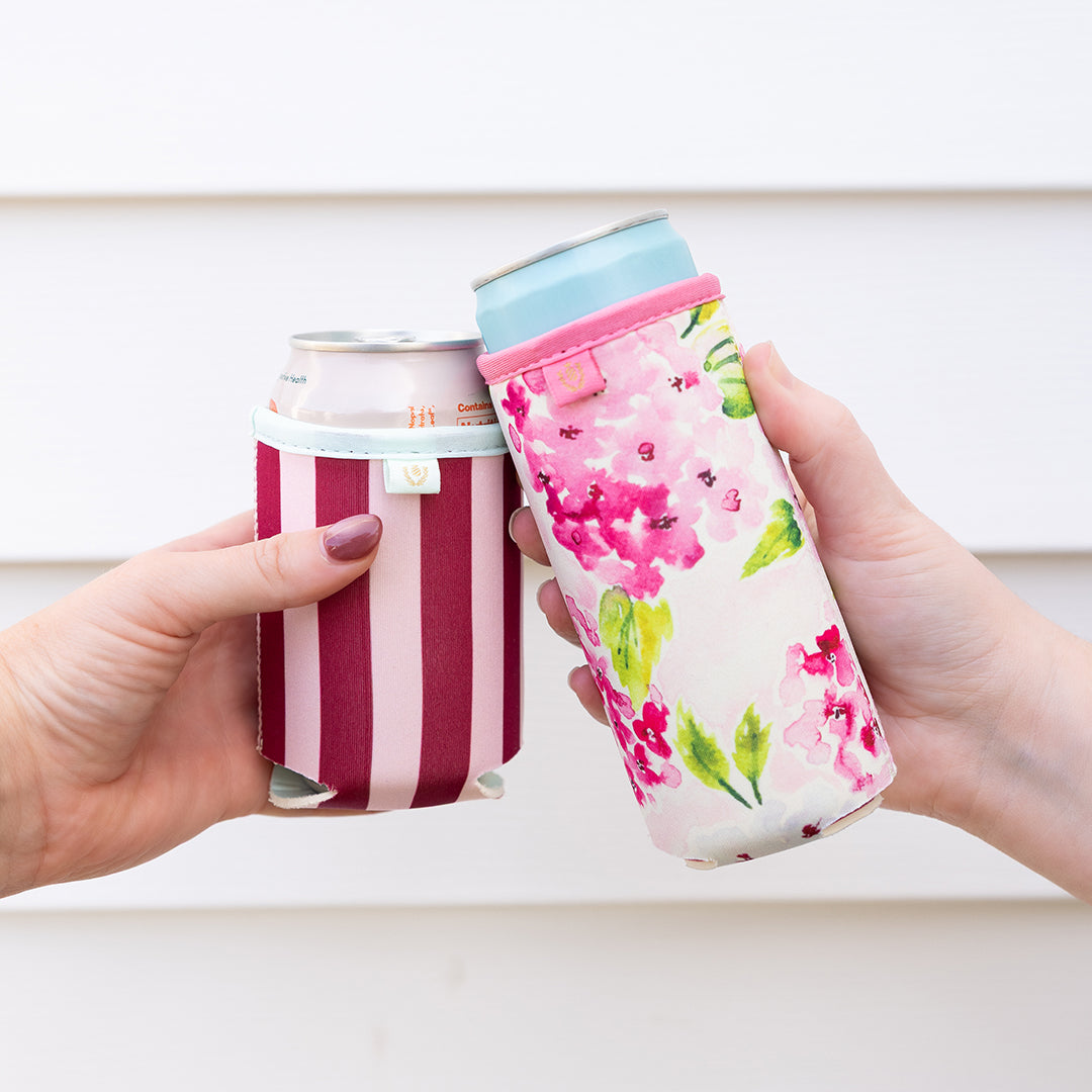 Two hands holding can coolers, one striped and one with floral design, against a white background.