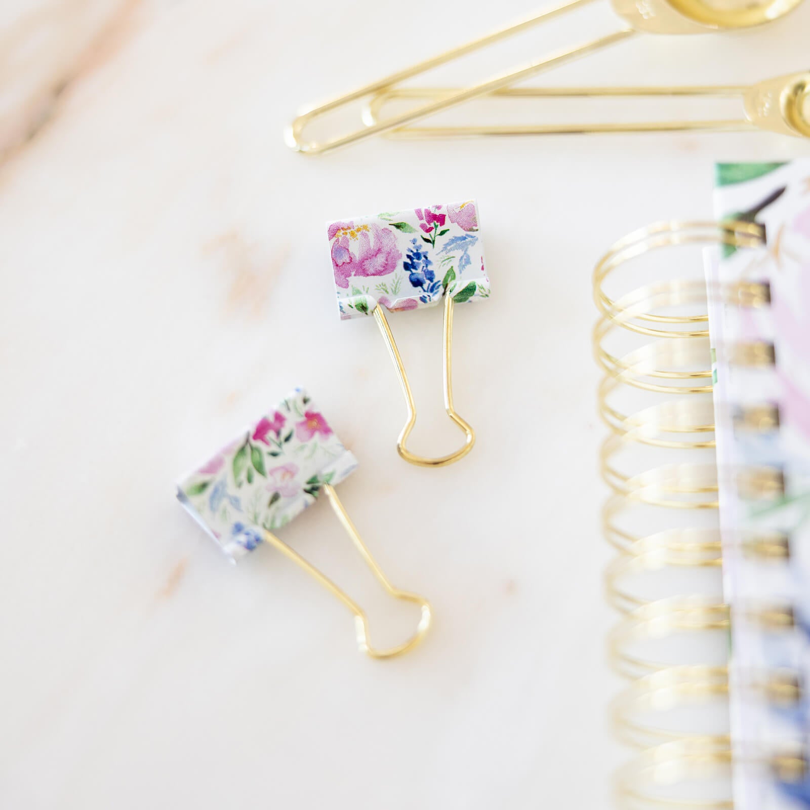 Two Simplified Binder Clips - Cottage Floral, featuring durable gold metal handles and a stylish floral pattern, rest beside the gold coil of a colorful notebook—perfect accents for an organized, chic workspace.