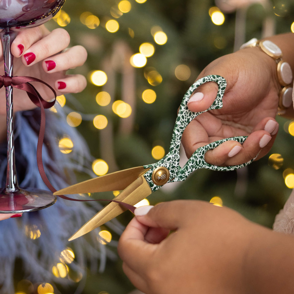 Emerald Floral Scissors with gold blades cutting burgundy ribbon tied to a wine glass, with a lit Christmas tree behind