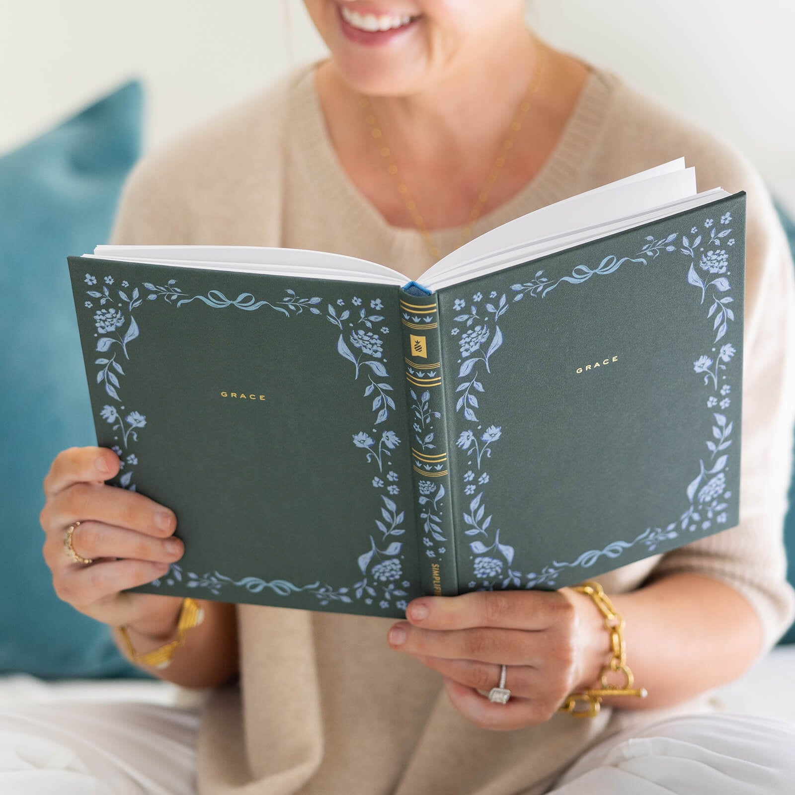 A woman in gold jewelry and a beige top smiles while reading the Simplified Linen Journal - Grace, featuring a green cover with blue hand-painted florals. She sits on a white surface with a teal pillow behind her.