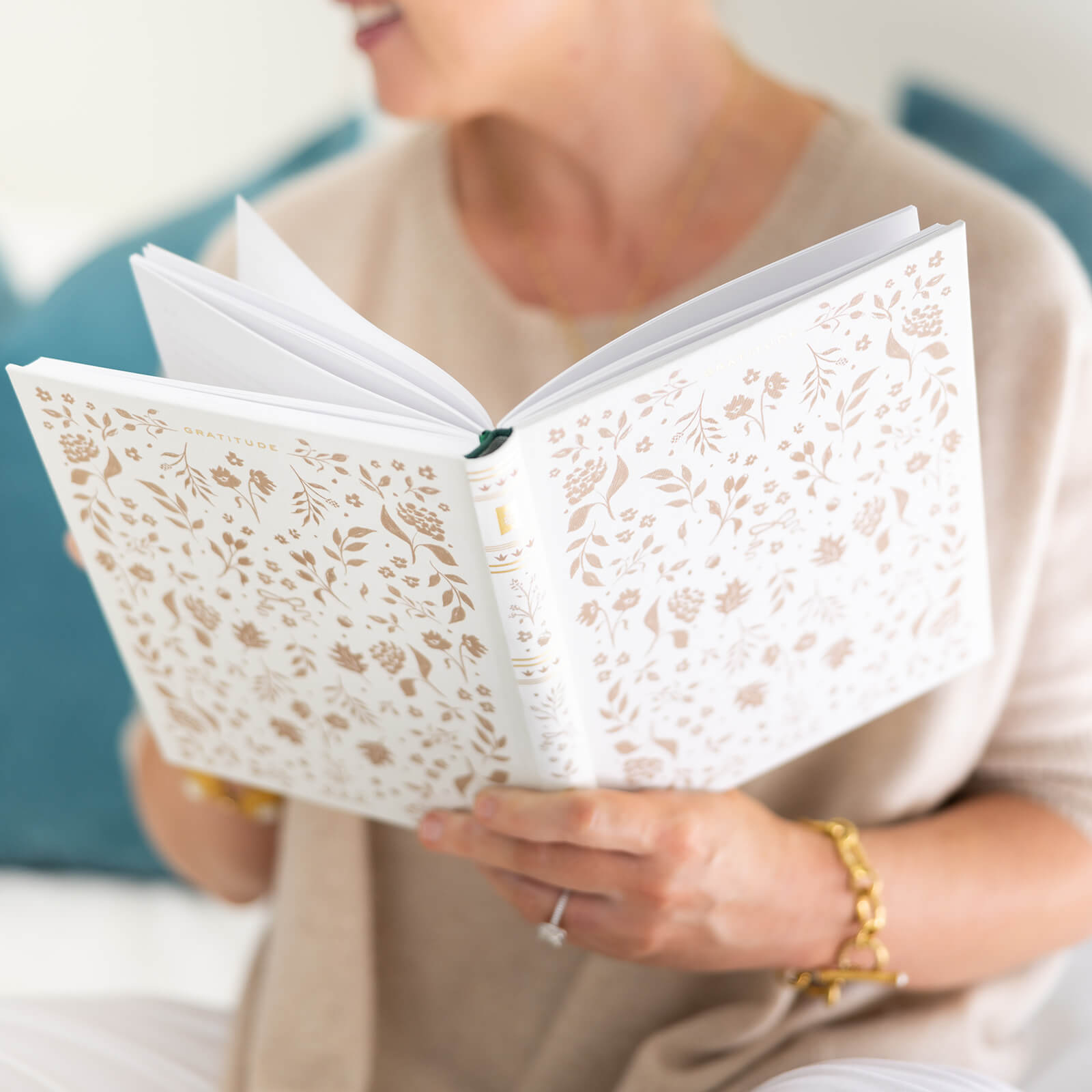 A person holds and reads the Simplified Linen Journal - Gratitude, featuring a linen hardcover with gold hand-painted details. Their upper body in a beige top, gold bracelet, and ring is visible against teal cushions in the background.