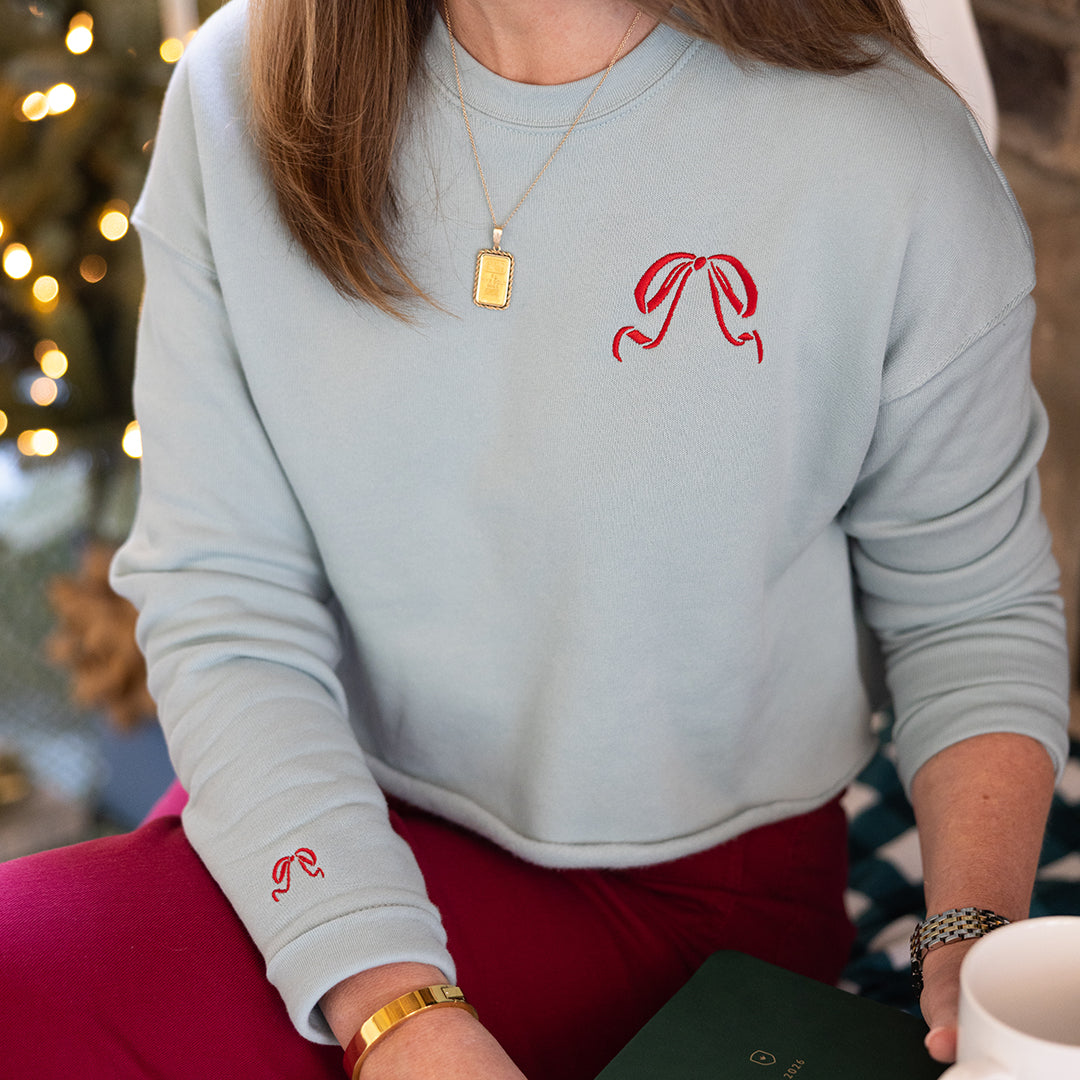 Someone in the Simplified Embroidered Crop Sweatshirt - Red Bow, paired with red pants and a gold necklace, holds a green book and white mug while seated near a lit Christmas tree.