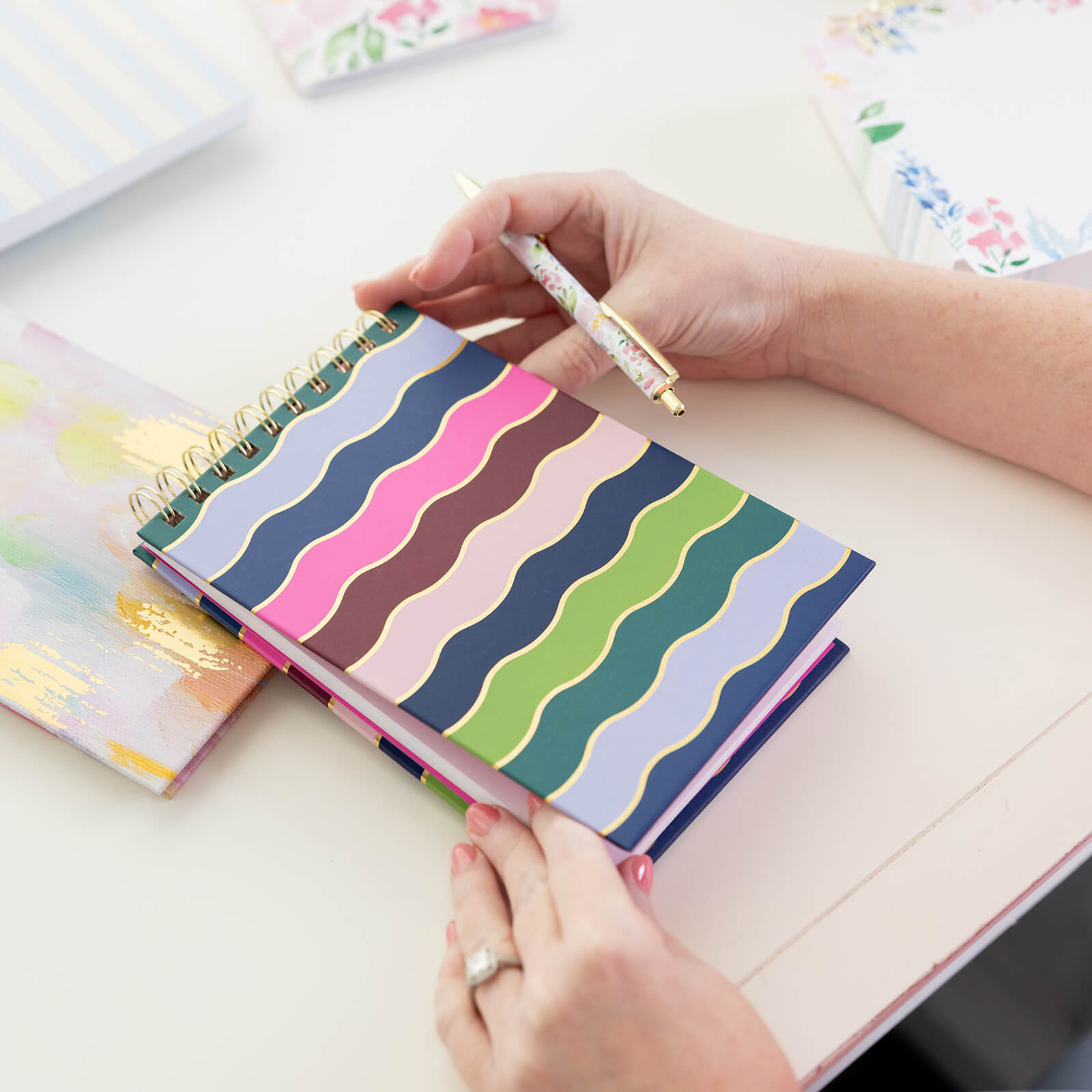 A person holds the Simplified Top Spiral Notebook - Rainbow Row, featuring colorful wavy stripes and paired with a floral pen, perfect for note-taking or gifting, while sitting at a desk with another pastel notebook beside them.