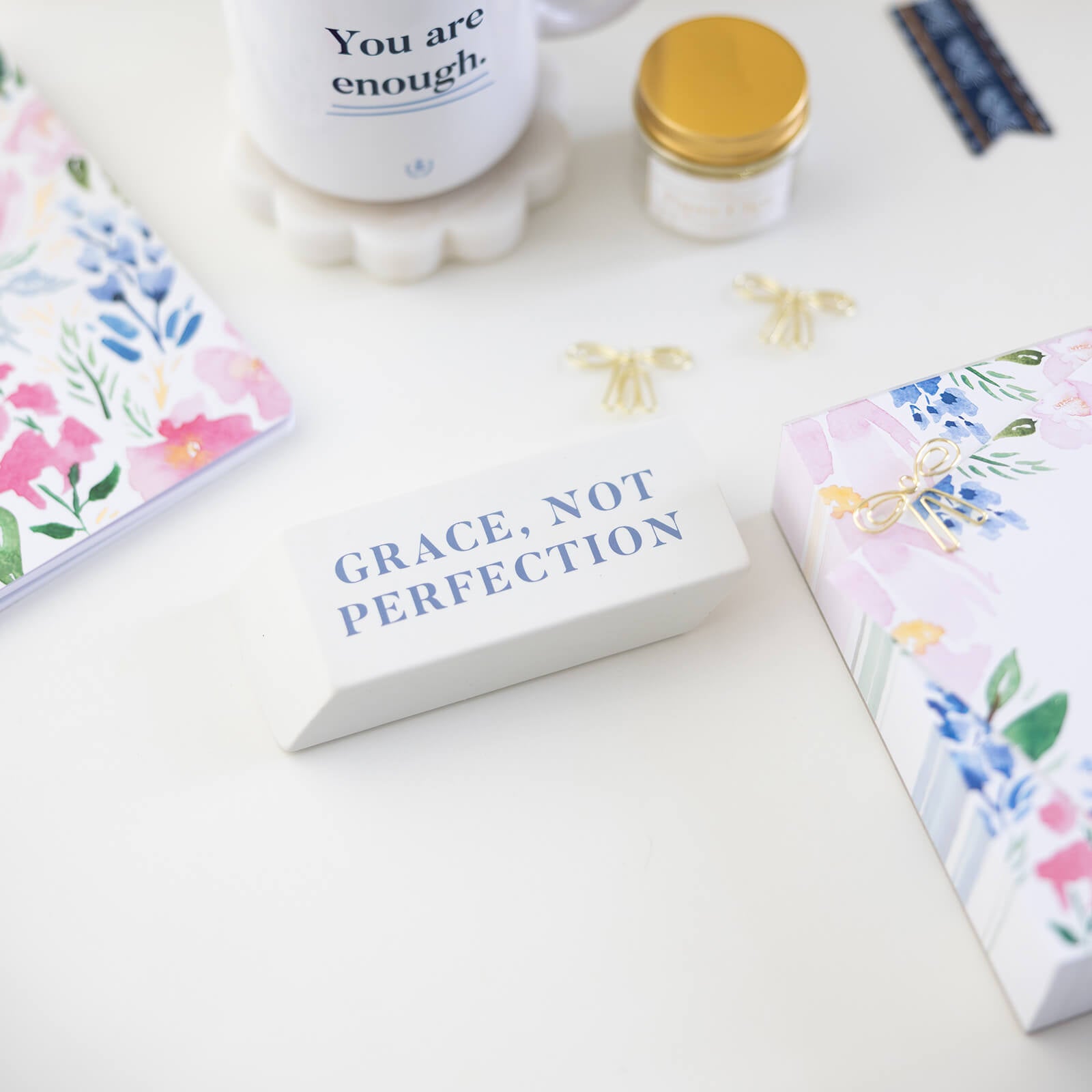 The Simplified Jumbo Eraser - Grace Not Perfection rests on a desk with floral notebooks, a You are enough mug, a gold-lidded jar, and gold paper clips—a charming accessory and thoughtful gift.