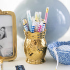 The Simplified Pineapple Pen Cup in gold holds vibrant checkered pens on a desk, beside a gold photo frame, blue bowl, white measuring tape, and a large blue balloon in the background.