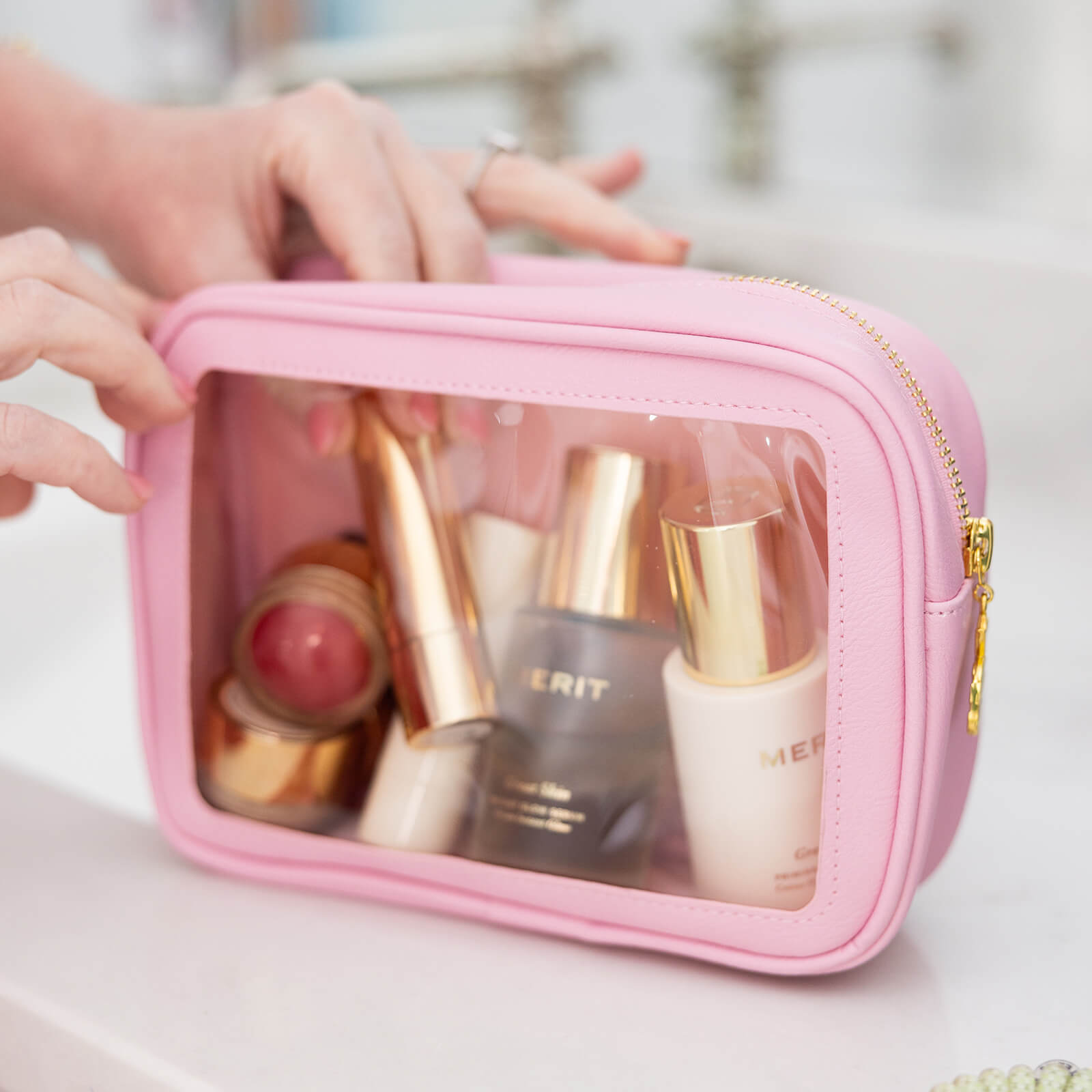 A person holds the Simplified Leatherette Travel Bag - Blush, featuring a pink zipper and clear window that displays neatly organized beauty products—ideal for travel essentials and TSA-friendly packing.
