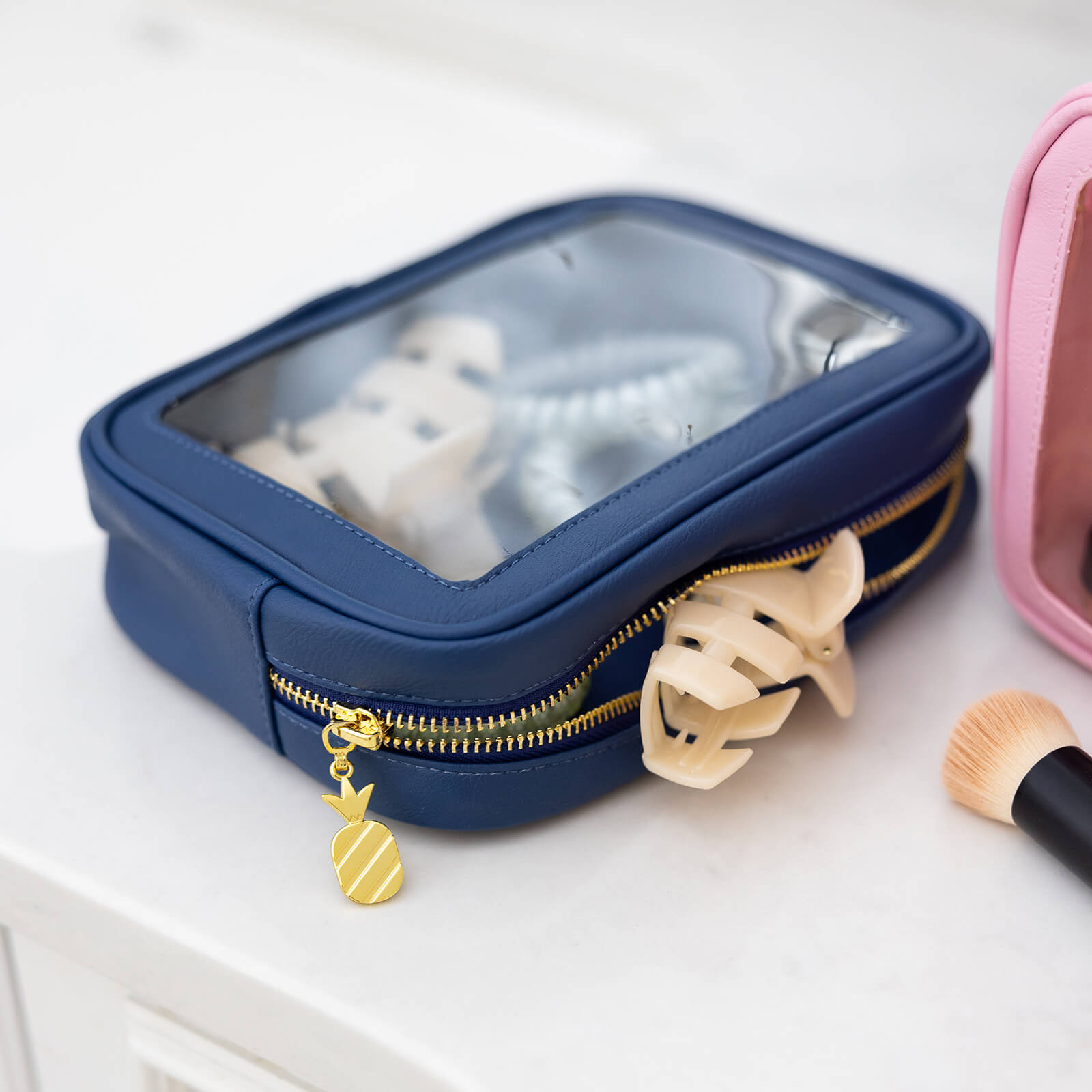 The Simplified Leatherette Travel Bag - Navy features a zippered clear top that reveals hair accessories inside. The pouch, shown on a white surface beside a makeup brush and pink bag, is perfect for organizing travel essentials.
