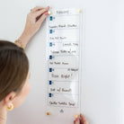 A woman mounts the Simplified Weekly Acrylic Wall Calendar on her wall, displaying February meals such as Crunchy Ranch Chicken, Egg Roll Bowls, Salmon Bites with rice, Pizza Night, and Chicken Tortilla Soup.