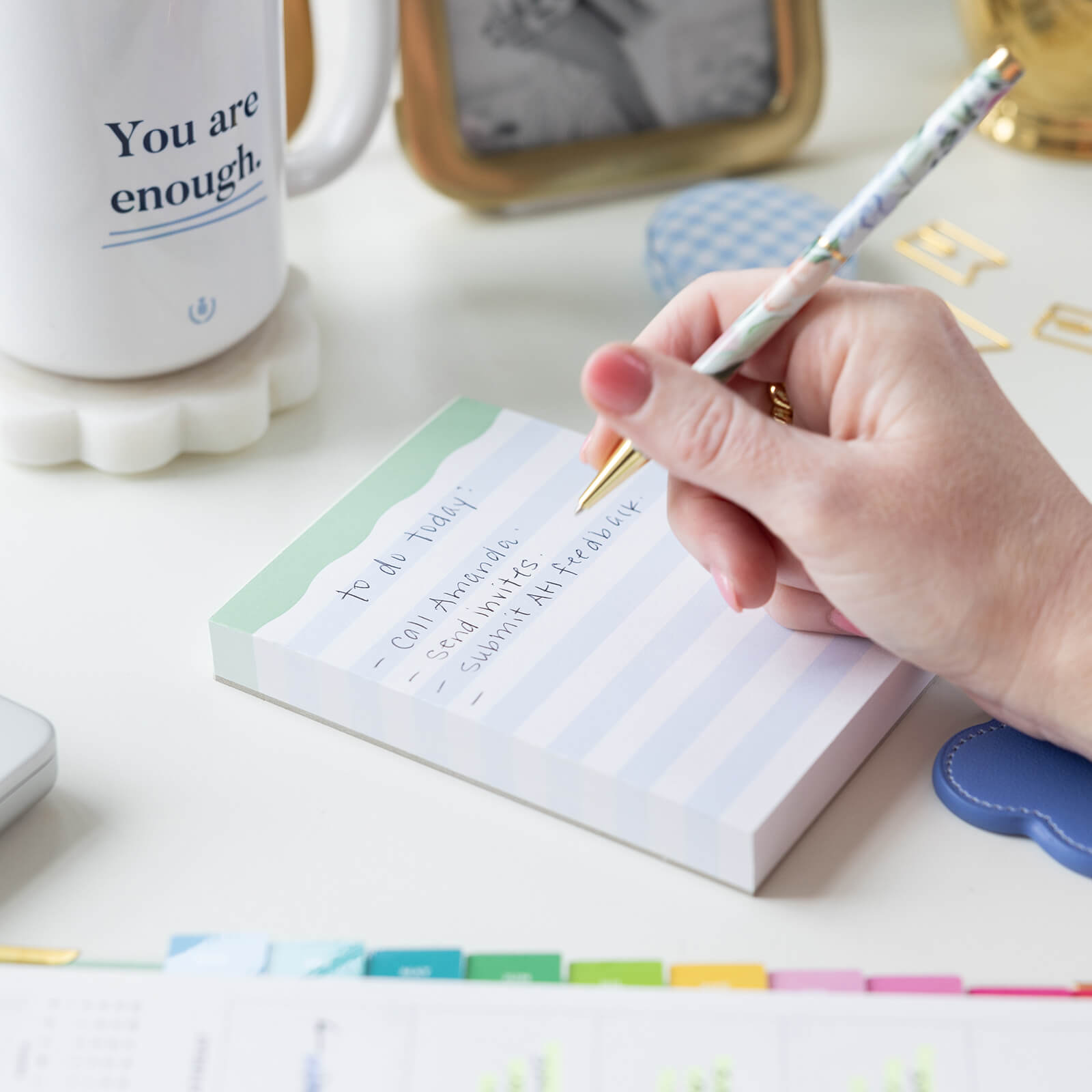 A hand writes notes on the Simplified Notepad - Blue Stripes, a true desk essential. Nearby sits a white mug reading You are enough, surrounded by colorful office supplies and a planner.