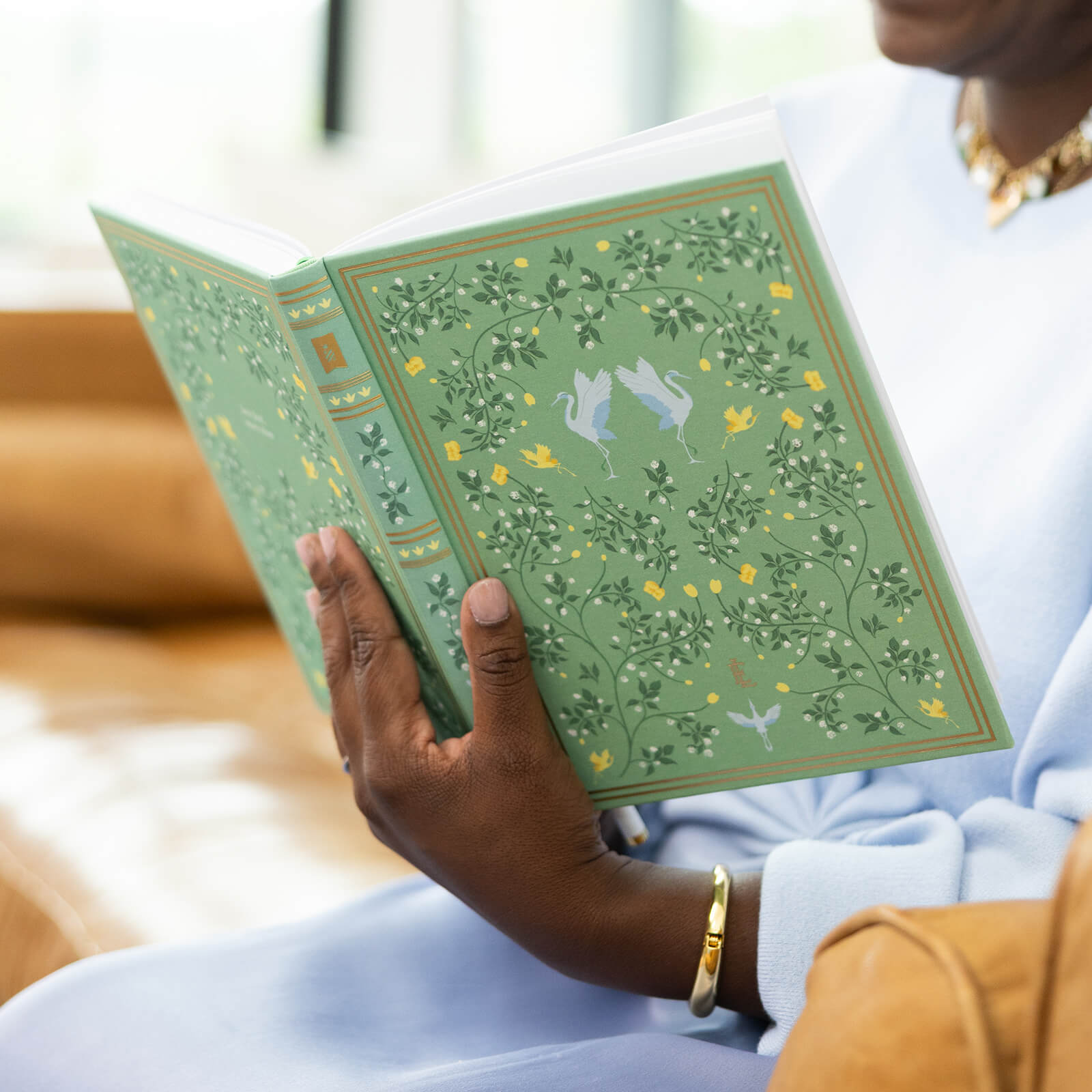 Seated on a tan couch in a light blue outfit with gold jewelry, a person reads the Simplified Linen Journal - Sage, adorned with ornate floral and bird designs—an ideal companion for creativity and reflection.