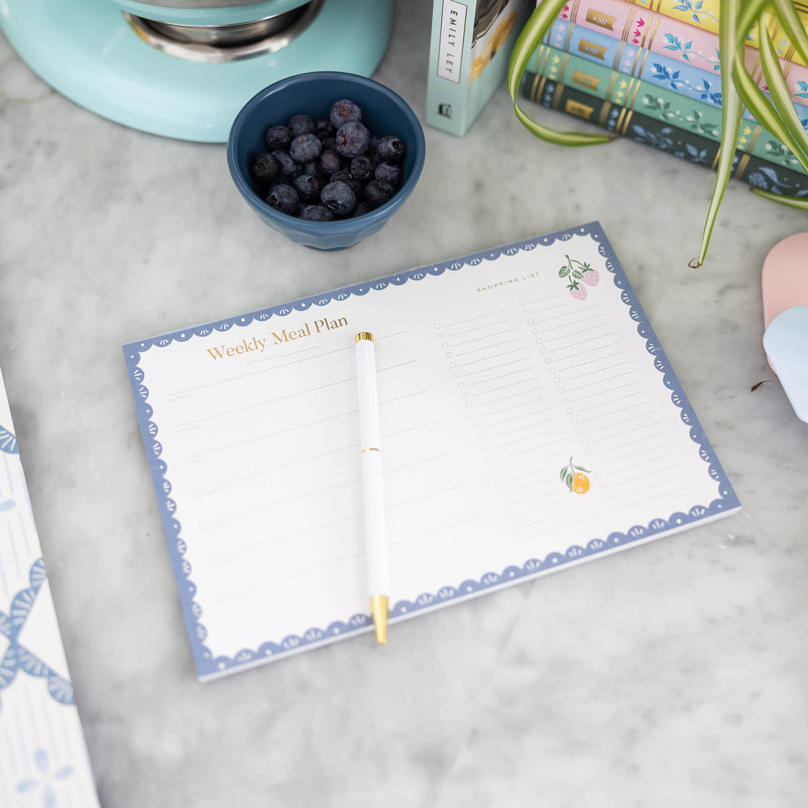 The Simplified Meal Planning Notepad with a pen rests on a marble surface, ready for planning meals and grocery lists, beside blueberries, books, and a potted plant.