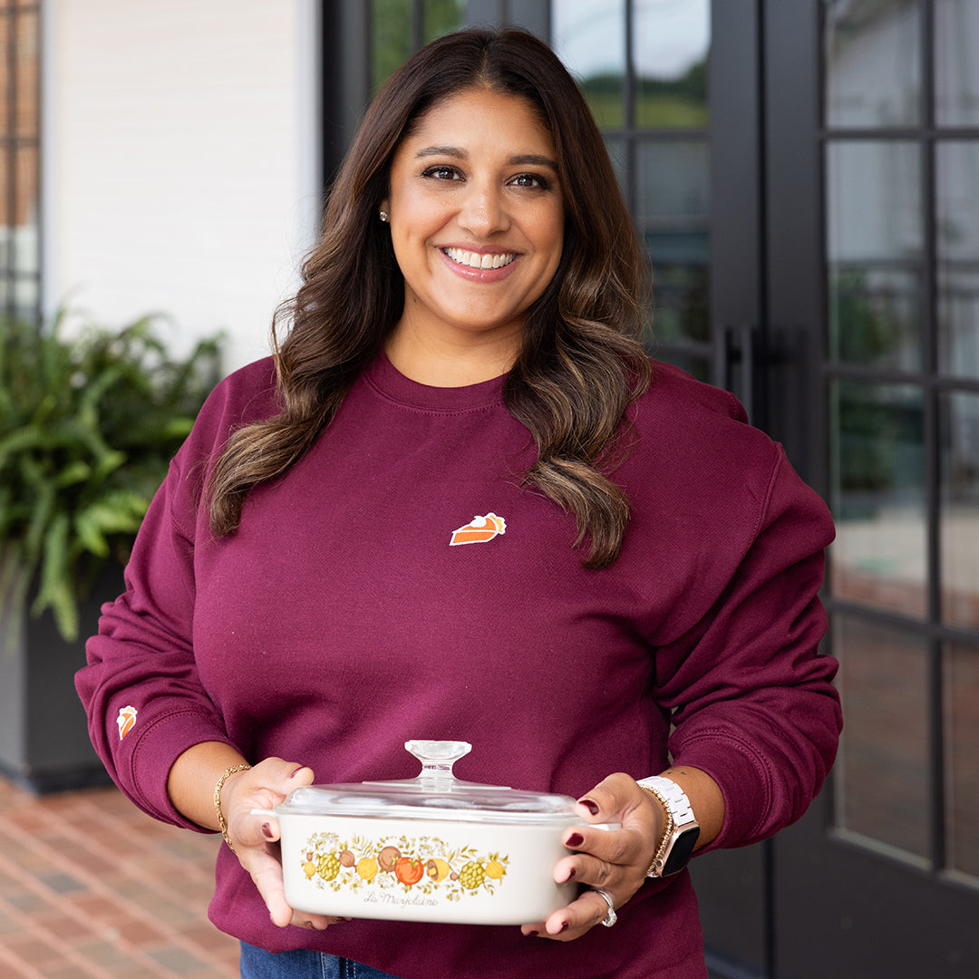 A woman with long dark hair smiles outdoors, wearing the Simplified Embroidered Crewneck - Sweet As Pie, which features an embroidered pie slice, and holds a vintage floral casserole dish in front of glass doors and green plants.
