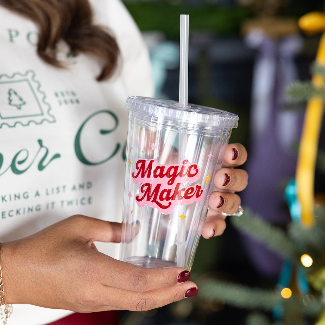 A person with dark red nails holds the Simplified Clear Tumbler - Magic Maker, a 16oz reusable cup with a straw, featuring "Magic Maker" in red with yellow and pink stars. A Christmas tree appears in the background.