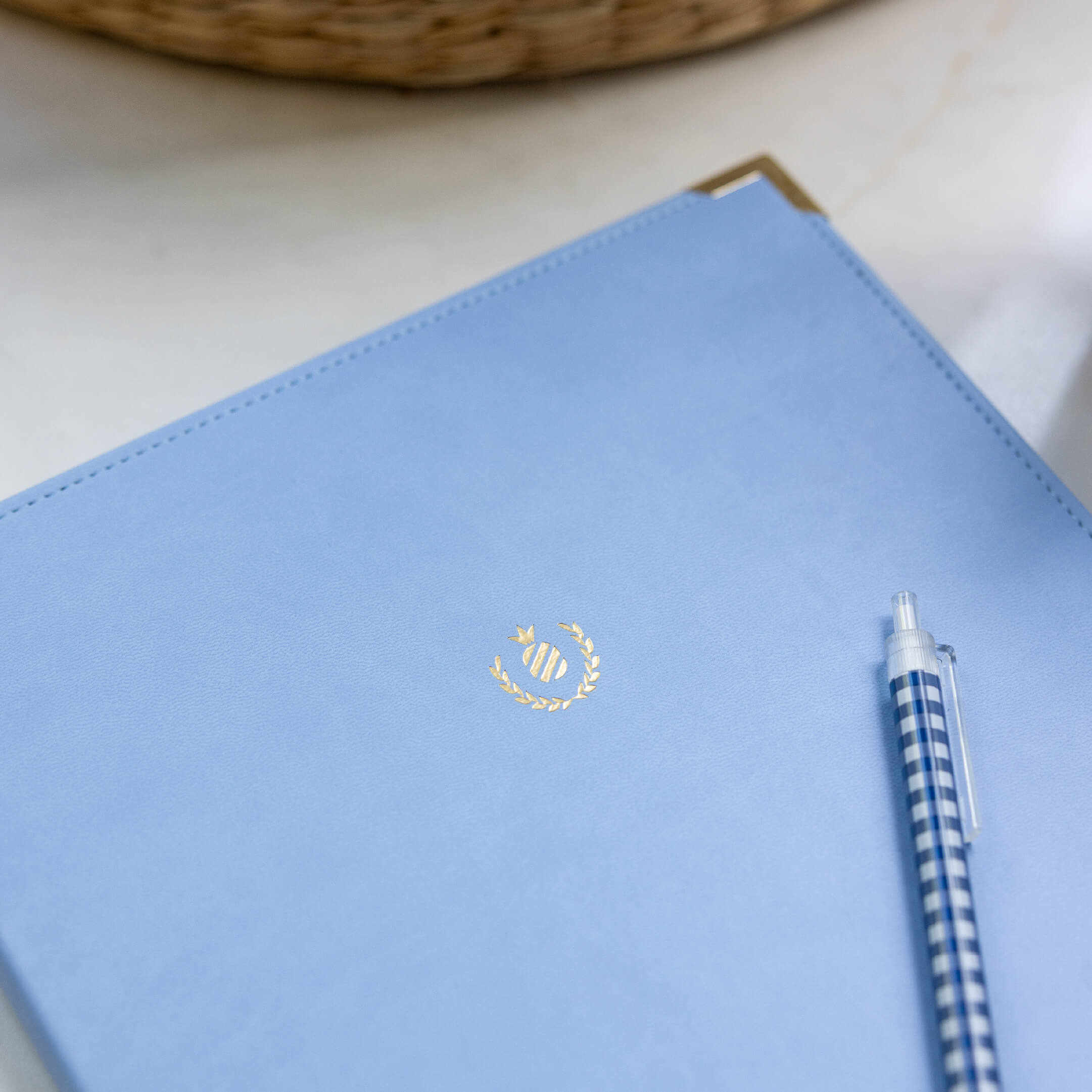The Simplified Leatherette Folio in French Blue, featuring a gold emblem, sits on a white surface next to a blue and white checkered pen. A woven basket appears in the background, creating an elegant workspace scene.