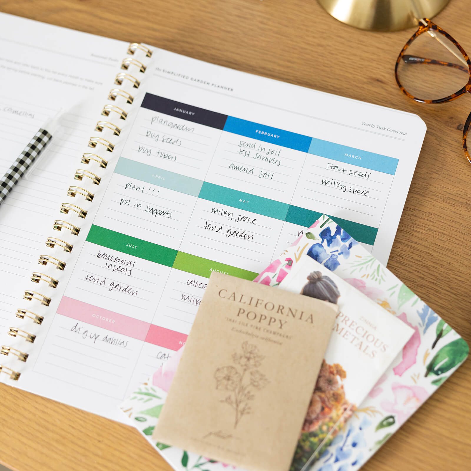 The Simplified Garden Planner, featuring handwritten notes, vibrant monthly calendars, a black-and-white pen, round eyeglasses, and assorted seed packets like “California Poppy,” is displayed on a wooden desk.