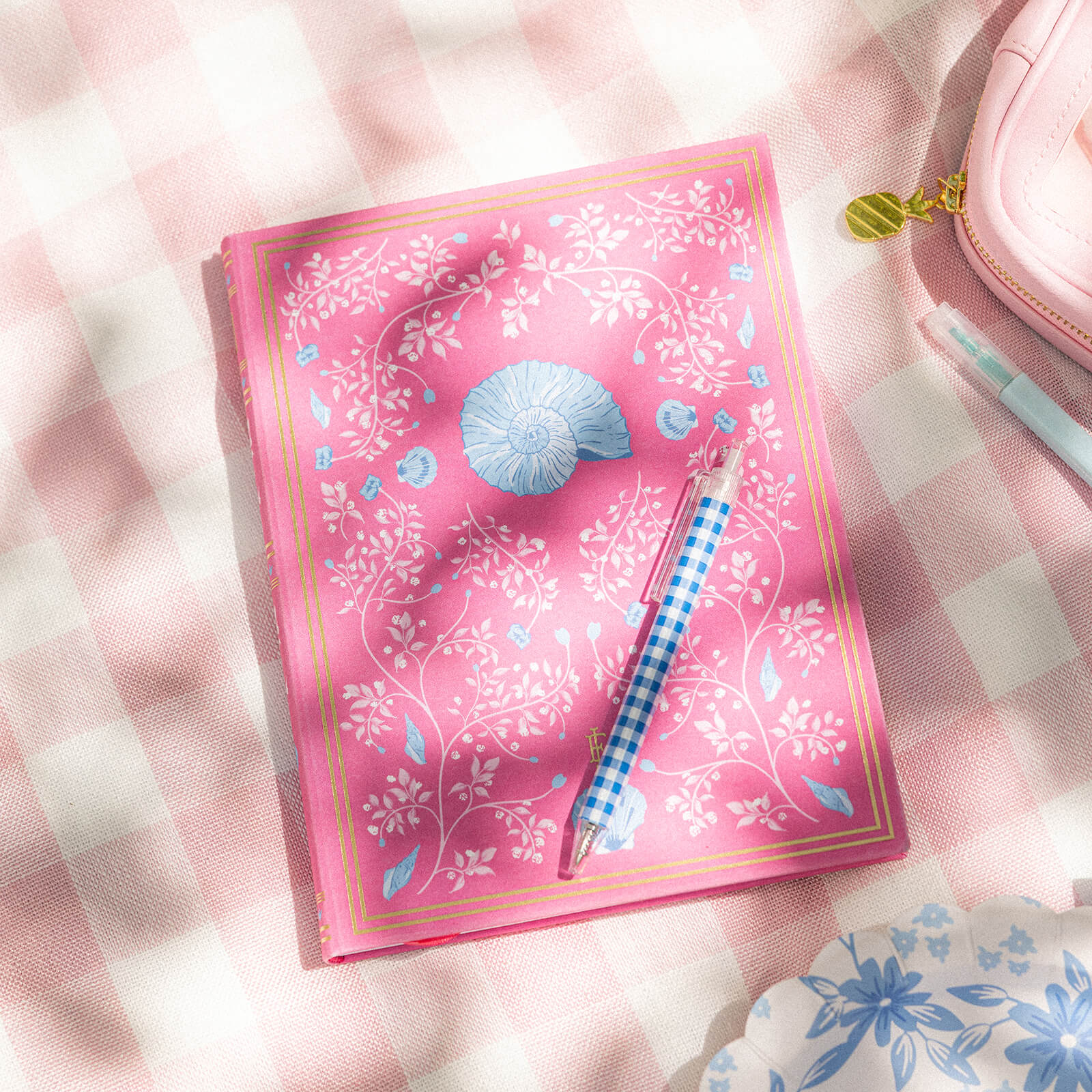 The Linen Journal - Raspberry by Simplified, featuring pink with white floral and blue shell designs, sits on a gingham cloth. A blue checkered pen rests atop it, beside a pink pouch and a plate with blue patterns.
