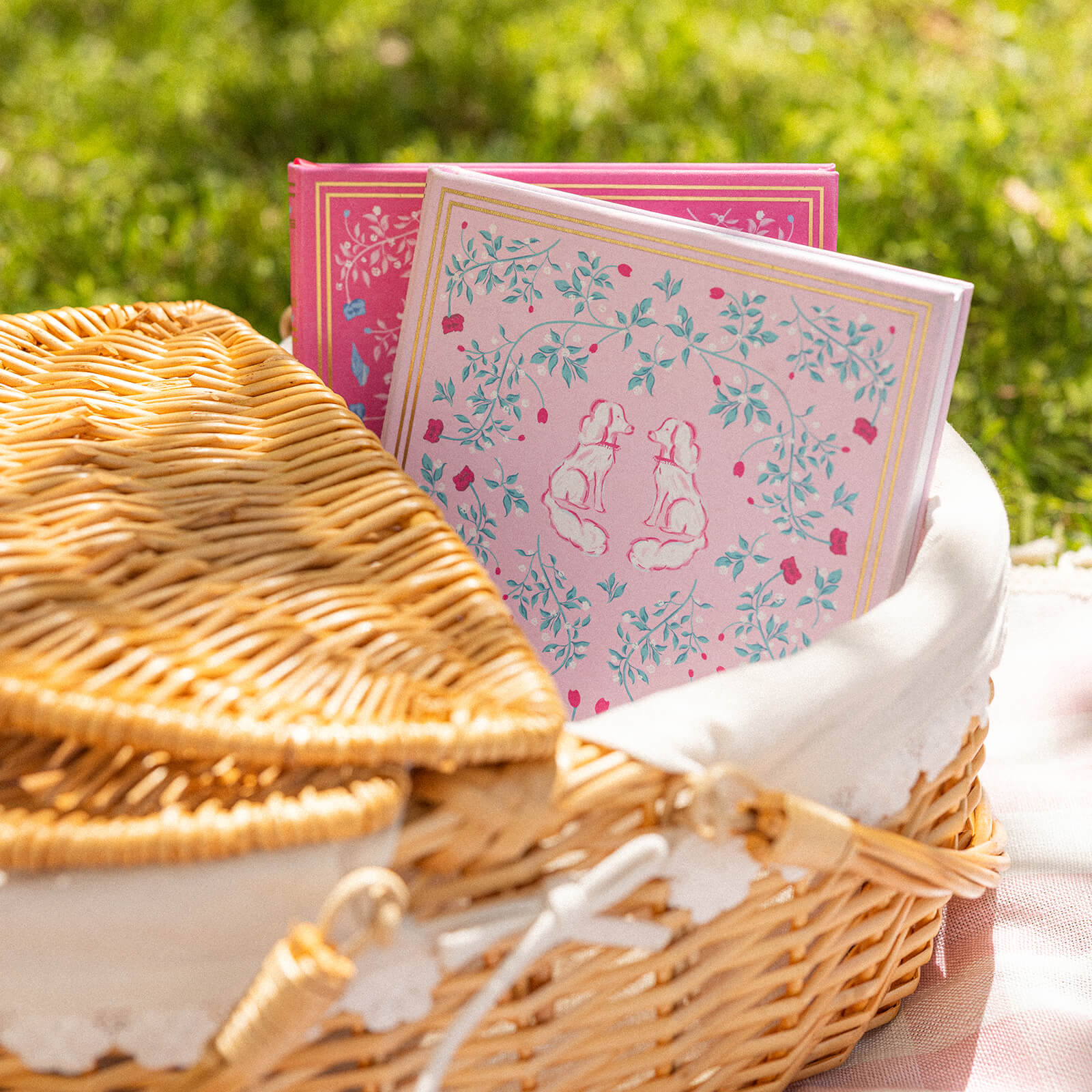 A wicker picnic basket holds two Simplified Linen Journal - Blush notebooks, inviting reflection and creativity. One displays white rabbits among flowers, resting on a pink-checked cloth in the grass.