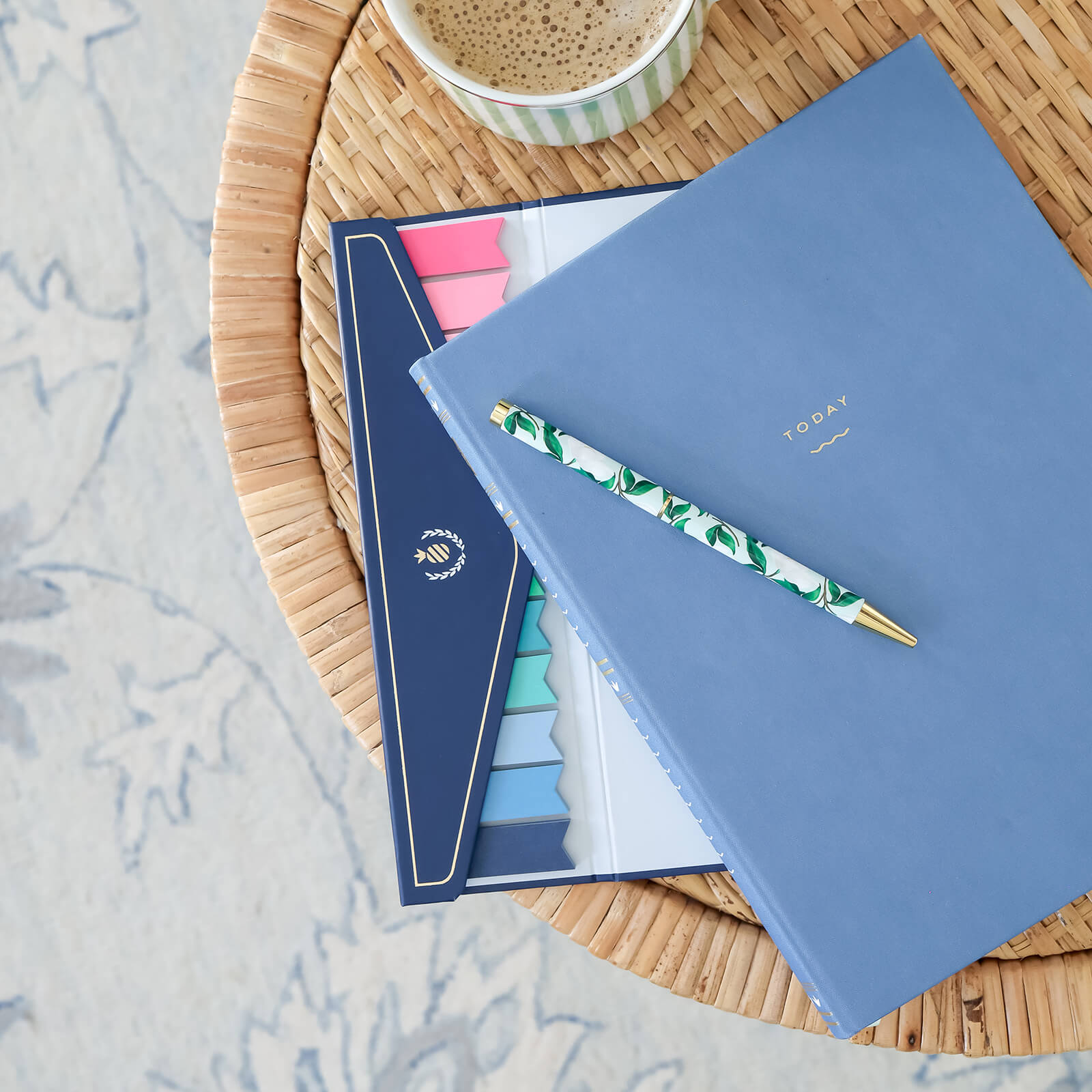 A Simplified Bullet Journal - French Blue with dot-grid pages labeled Today, a floral pen, colored sticky tabs, and a coffee cup sit on a woven table, with a patterned rug partially visible in the background.
