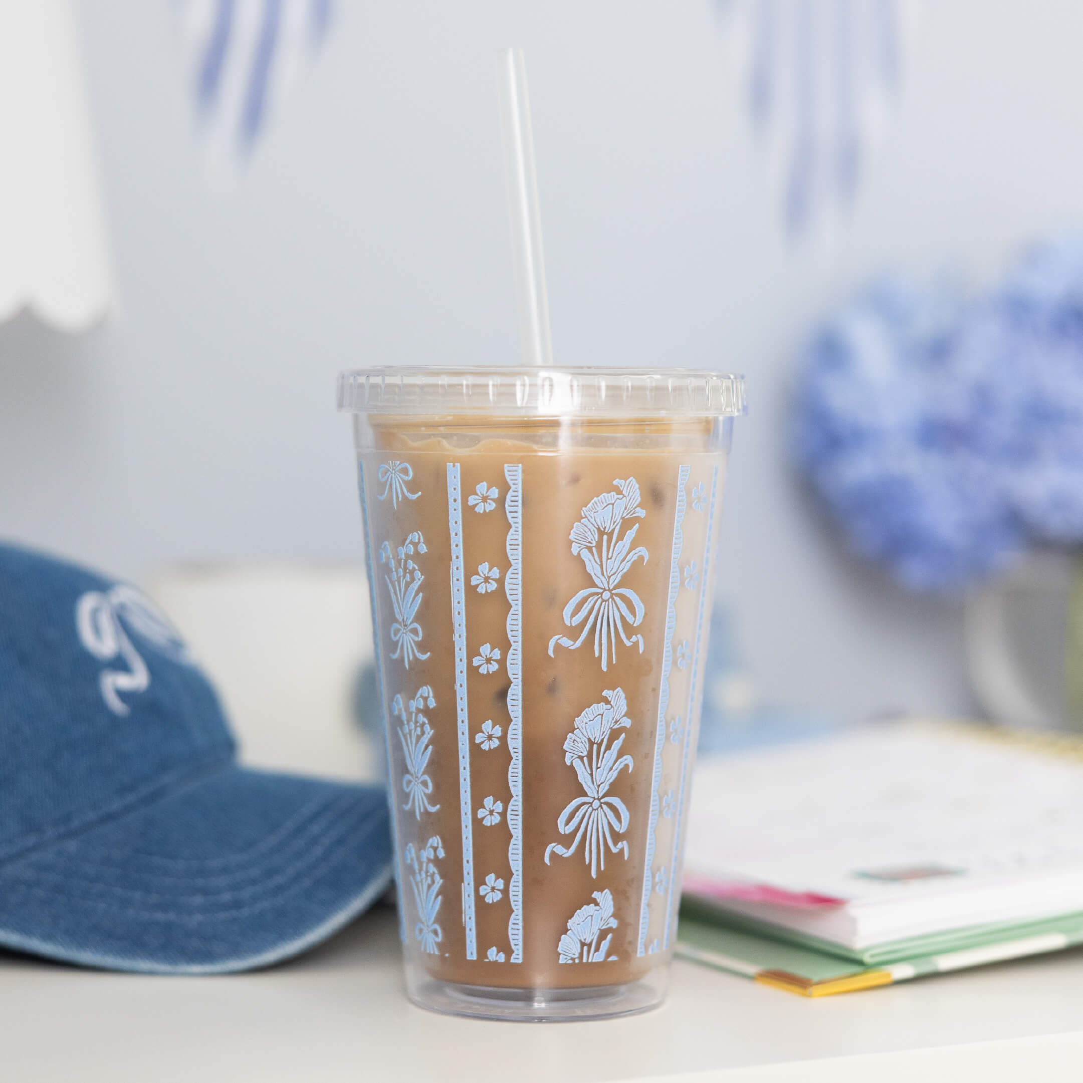 The Simplified Clear Tumbler - Poppy Posies, a 16oz reusable cup with blue and white patterns, holds iced coffee with a clear straw. It sits on a desk next to a blue hat, books, and blurred blue flowers in the background.