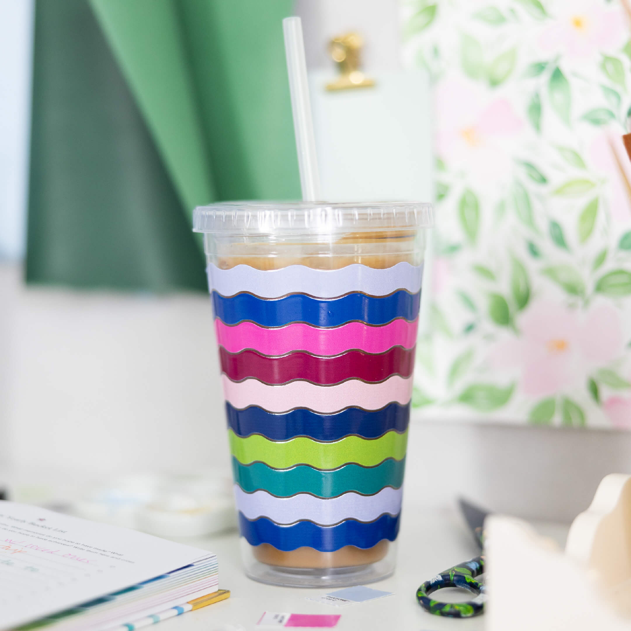 The Simplified Clear Tumbler - Rainbow Row holds 16oz, comes with a lid and straw, and features blue, pink, green, and purple wavy stripes. It sits on a white desk alongside books and floral decor.
