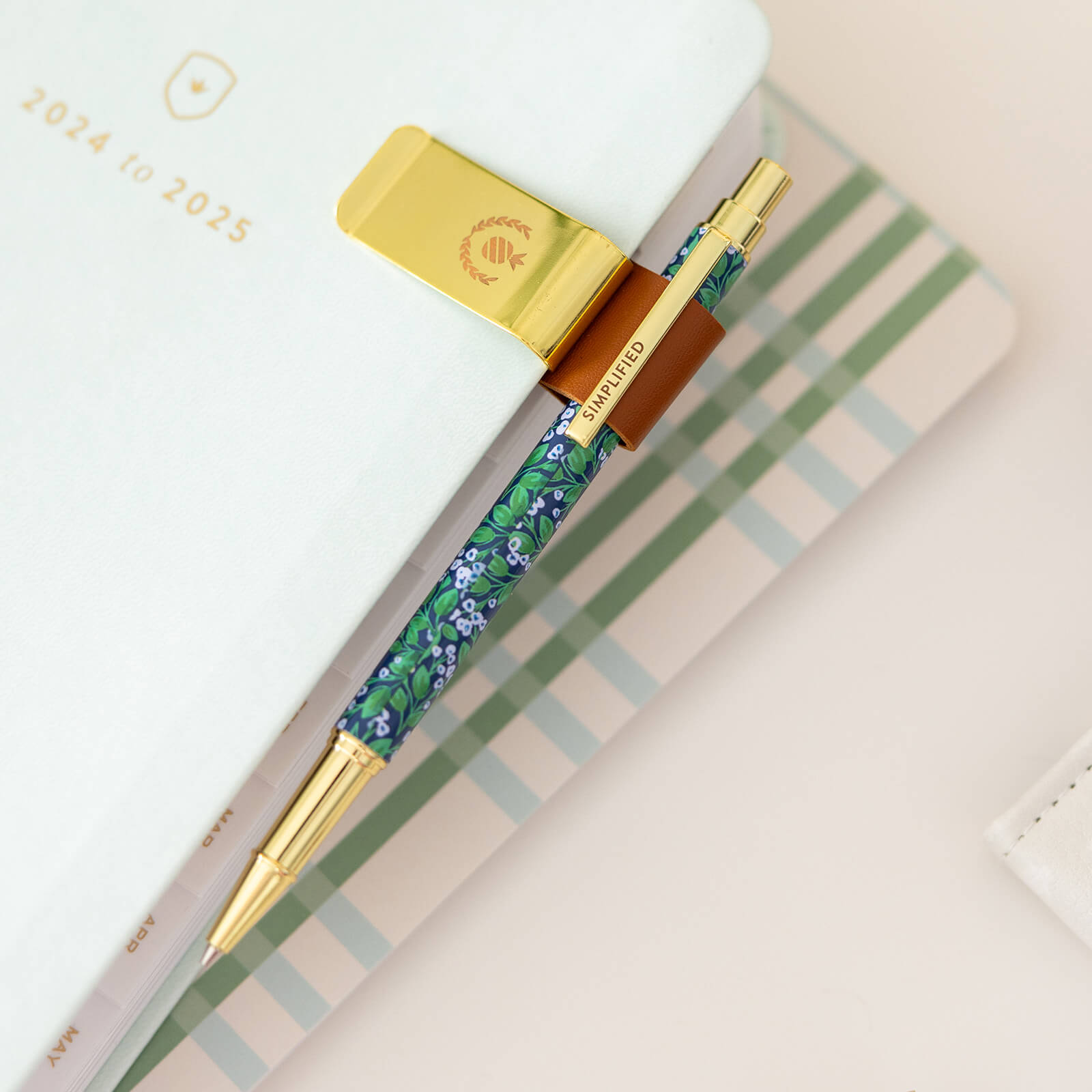 A 2024-2025 planner with a gold clip holds a floral pen with gold accents, resting on a striped desk beside Simplifieds Misty Meadow mechanical pencil featuring a sleek metal body.