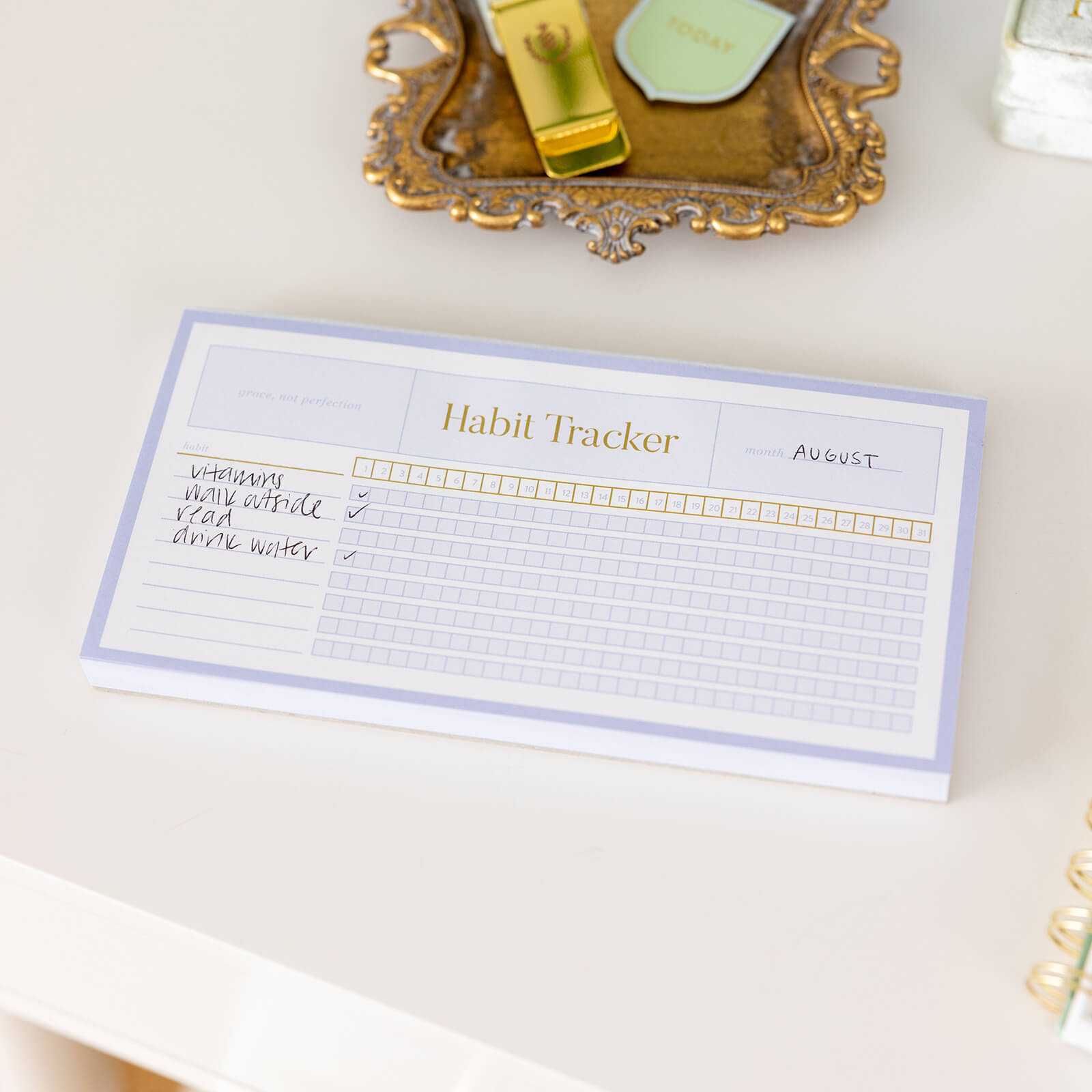 The Simplified Notepad - Habit Tracker for August tracks habits like vitamins, walking outside, reading, and drinking water with checkboxes. Grocery list and reminder notepads rest on a decorative tray amid desk accessories.