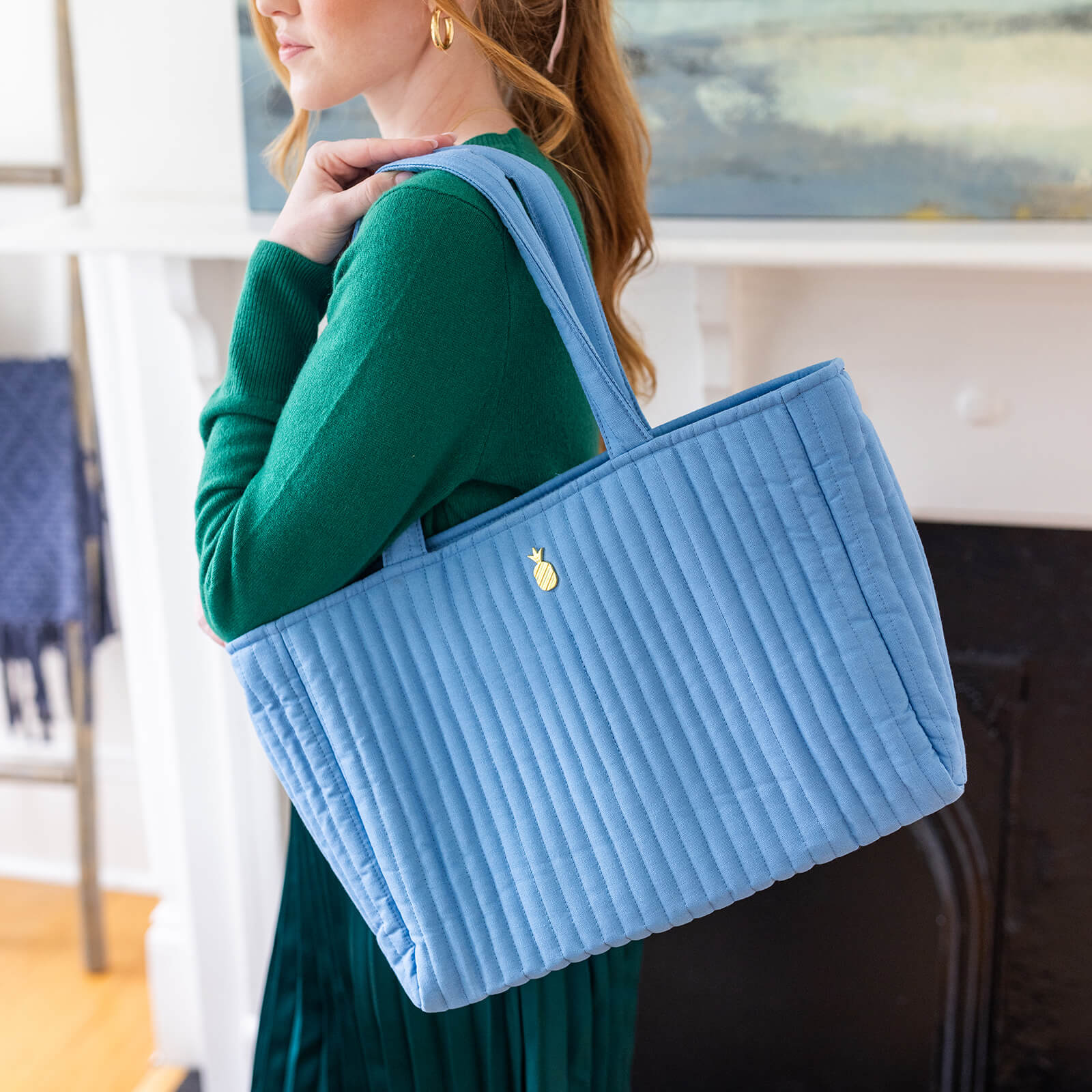 A woman in a green long-sleeve dress carries the Simplified Quilted Tote Bag in French Blue, featuring padded handles and a gold pineapple charm.