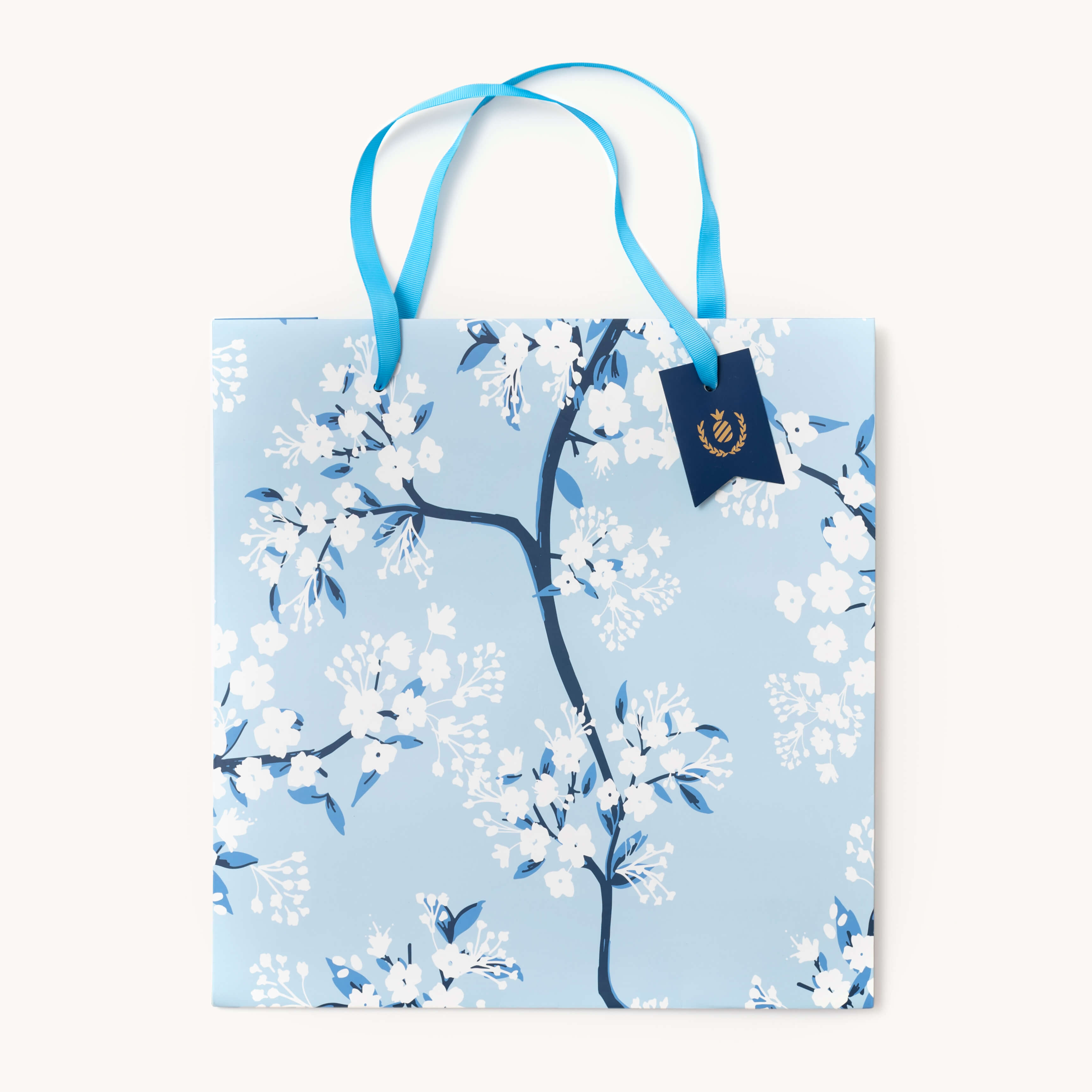 The Simplified Gift Bag - Cherry Blossoms features a light blue design with blue handles, white and blue blossom branches, a sturdy build, dark blue tag with gold emblem, and comes in various sizes and designs.