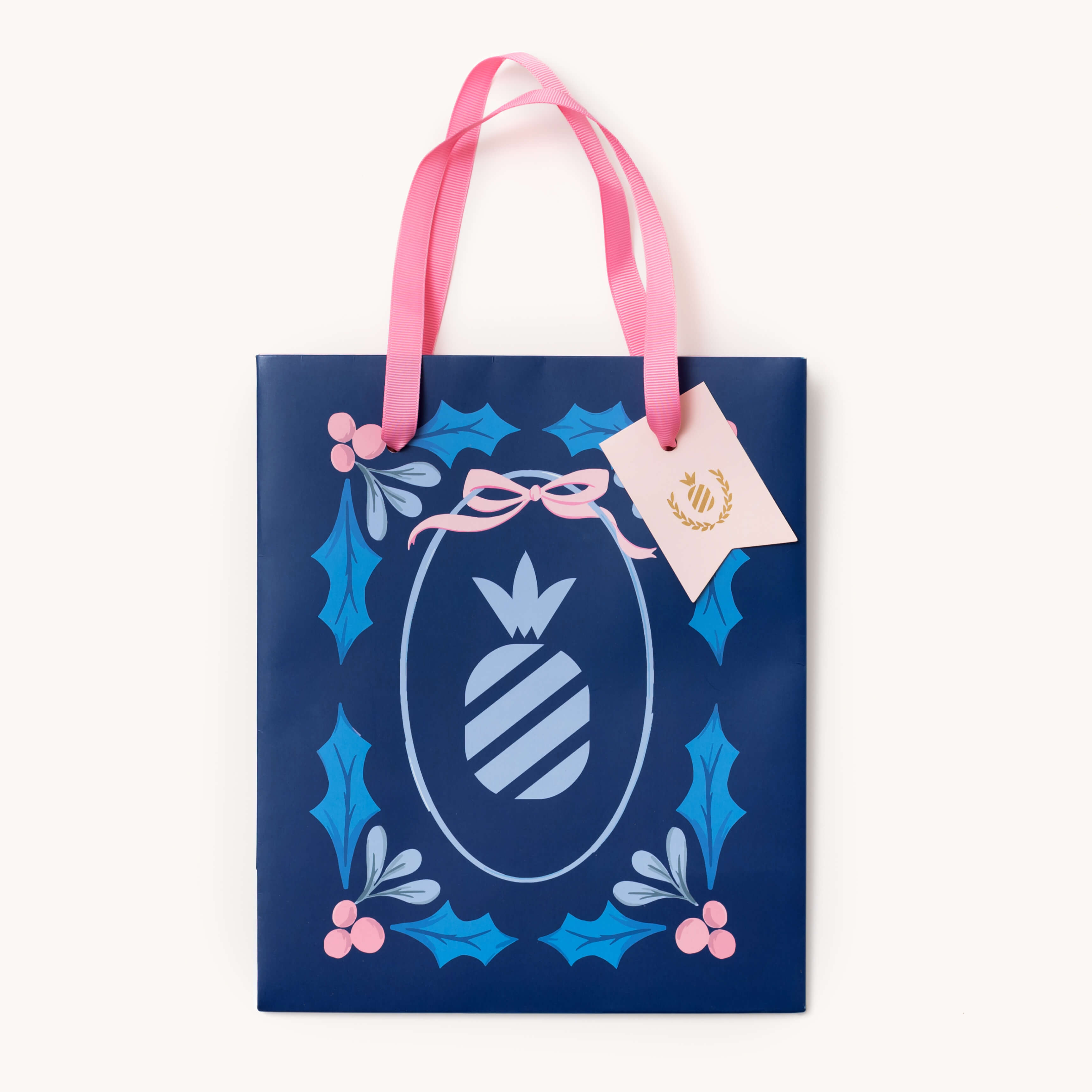 The Simplified Gift Bag - Navy Pineapple Stamp features pink handles, a sturdy build, and a festive pineapple stamp design. Includes a cream tag with gold wreath and is available in multiple sizes.