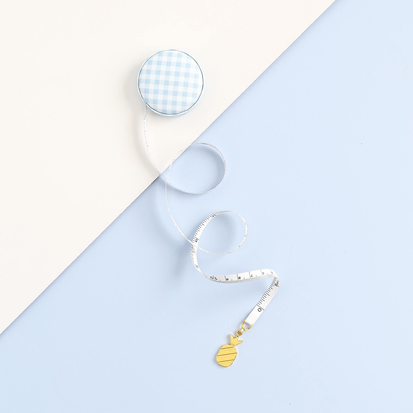 A Simplified Measuring Tape in Carolina Gingham blue and white rests on a light background, partially extended to reveal a small gold pineapple charm at the end.