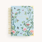 The 2026 Spiral Daily Simplified Planner - Faraway Land by Simplified features a light blue, spiral-bound cover with hand-painted pink, green, and black floral and butterfly designs, gold Simplified Planner text, and gold metal corner accents.