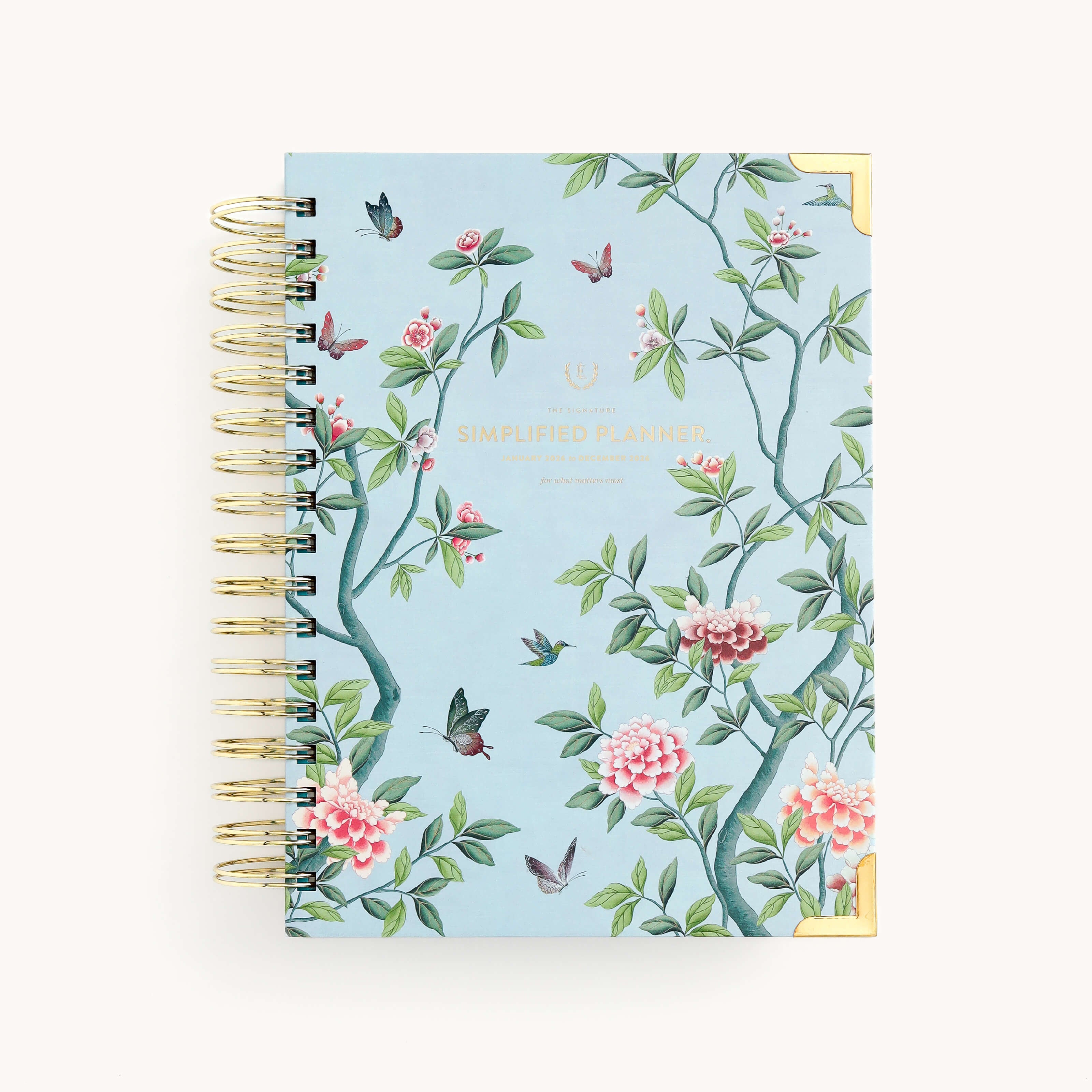 The 2026 Spiral Daily Simplified Planner - Faraway Land by Simplified features a light blue, spiral-bound cover with hand-painted pink, green, and black floral and butterfly designs, gold Simplified Planner text, and gold metal corner accents.