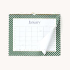 The 2026 Wall Calendar by Simplified features a green and white checkered border and is displayed open to January, with a partially turned page showing the next month beneath, hung on a white wall.