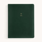 The Simplified 2026 Bookbound Weekly Dapperdesk Planner in Evergreen has a dark green leatherette cover with rounded corners, topped with a gold embossed shield and 2026. Set against an off-white background, it adds style to planning.