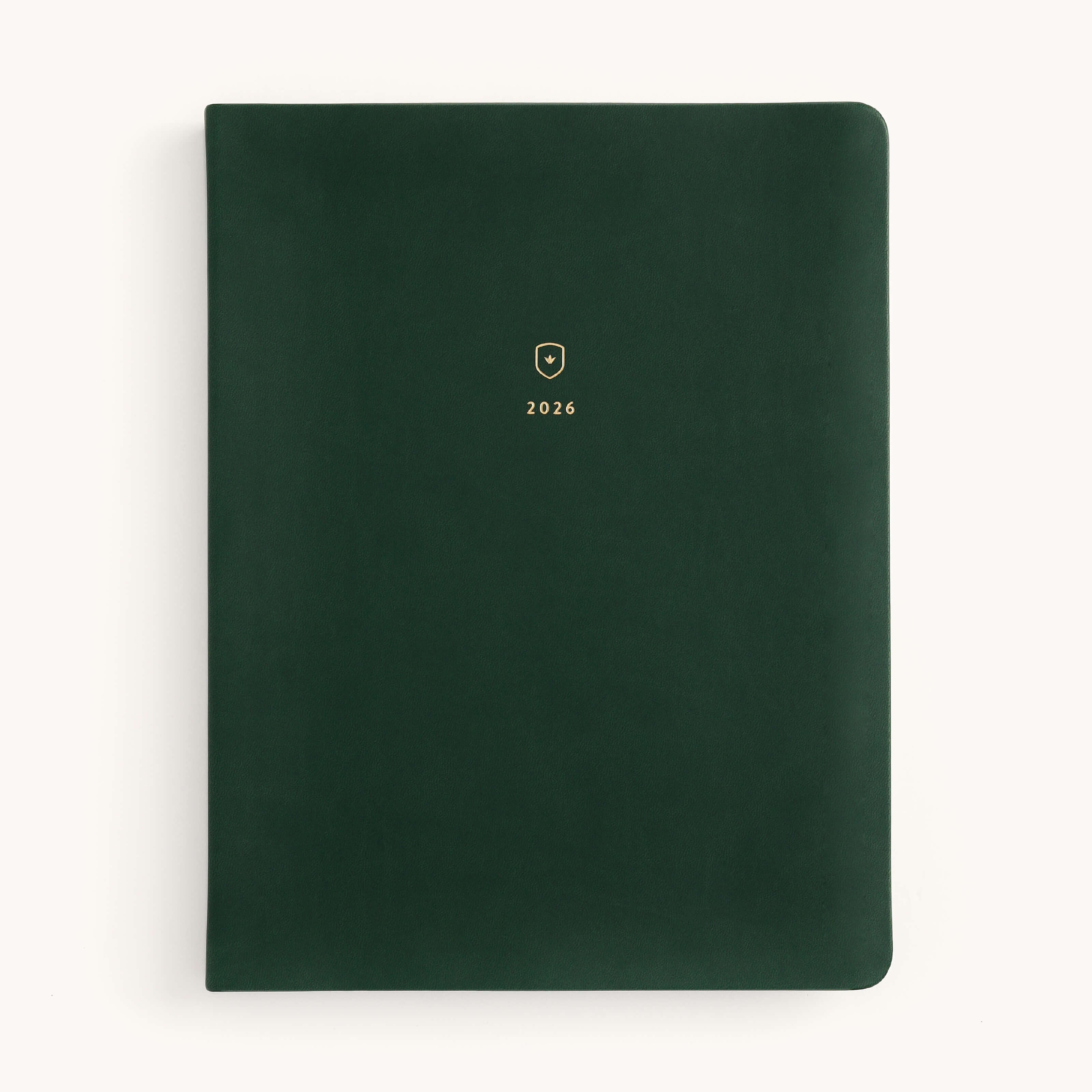 The Simplified 2026 Bookbound Weekly Dapperdesk Planner in Evergreen has a dark green leatherette cover with rounded corners, topped with a gold embossed shield and 2026. Set against an off-white background, it adds style to planning.