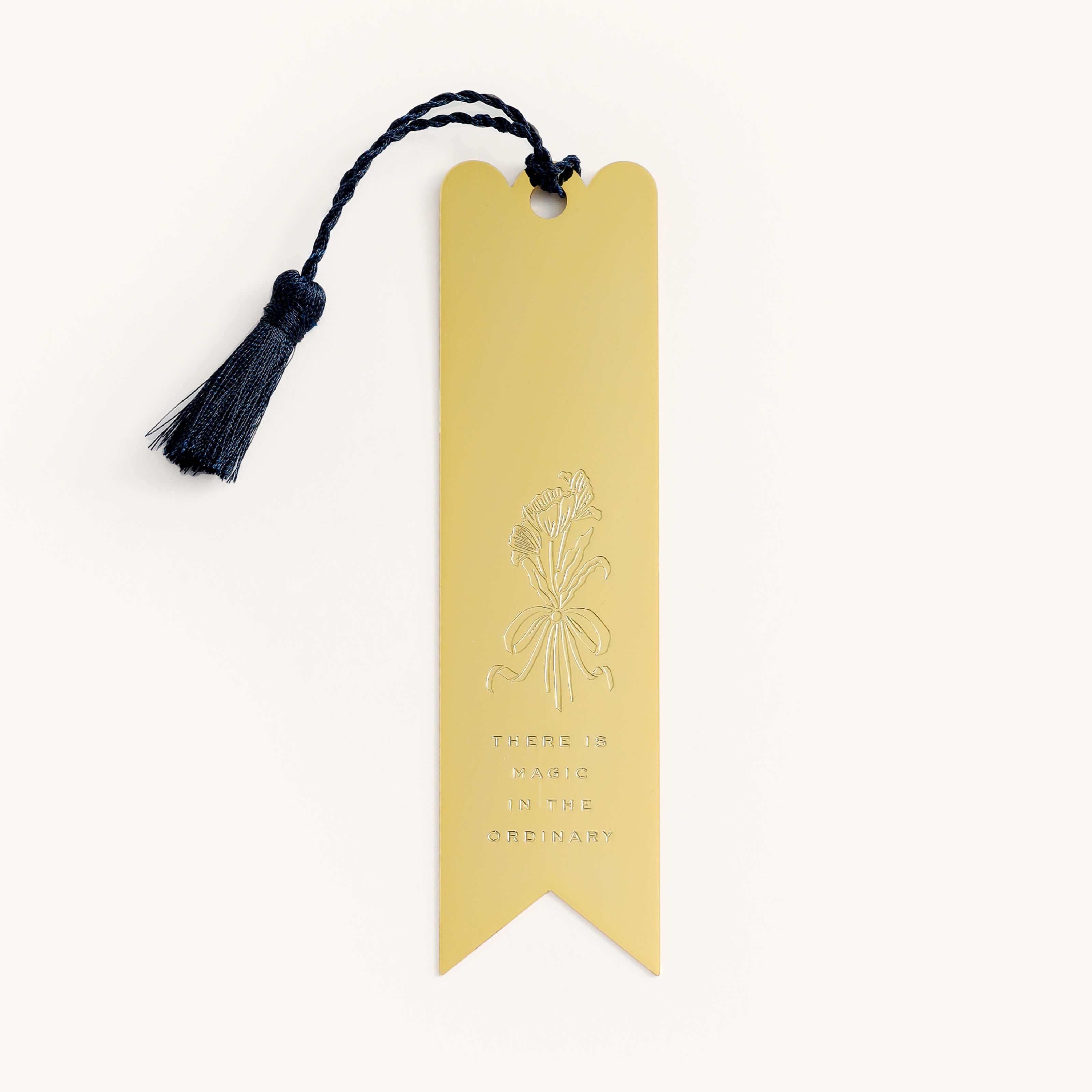 The Simplified Engraved Bookmark - Magic In The Ordinary features a gold metal design with a navy blue tassel, floral engraving, and the phrase “There is magic in the ordinary” below the illustration.