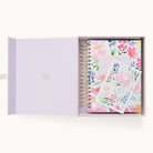 An open box with a light purple interior holds the 2026 Spiral Daily Simplified Planner - Cottage Floral by Simplified and a matching floral notecard, both adorned with colorful pink, blue, and green flowers.