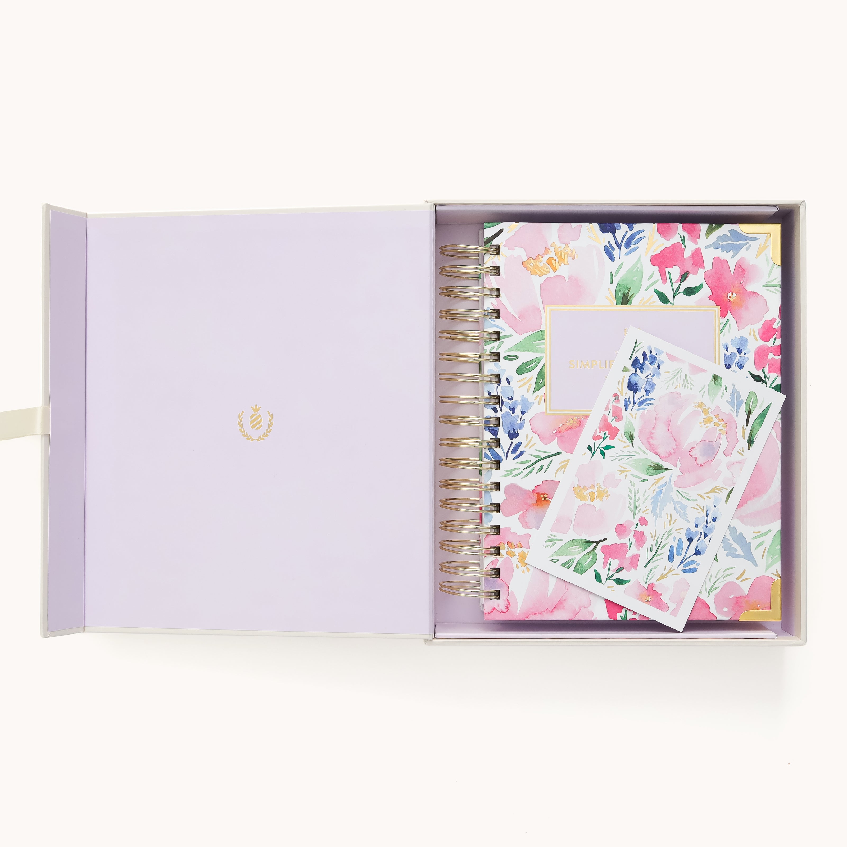 An open box with a light purple interior holds the 2026 Spiral Daily Simplified Planner - Cottage Floral by Simplified and a matching floral notecard, both adorned with colorful pink, blue, and green flowers.