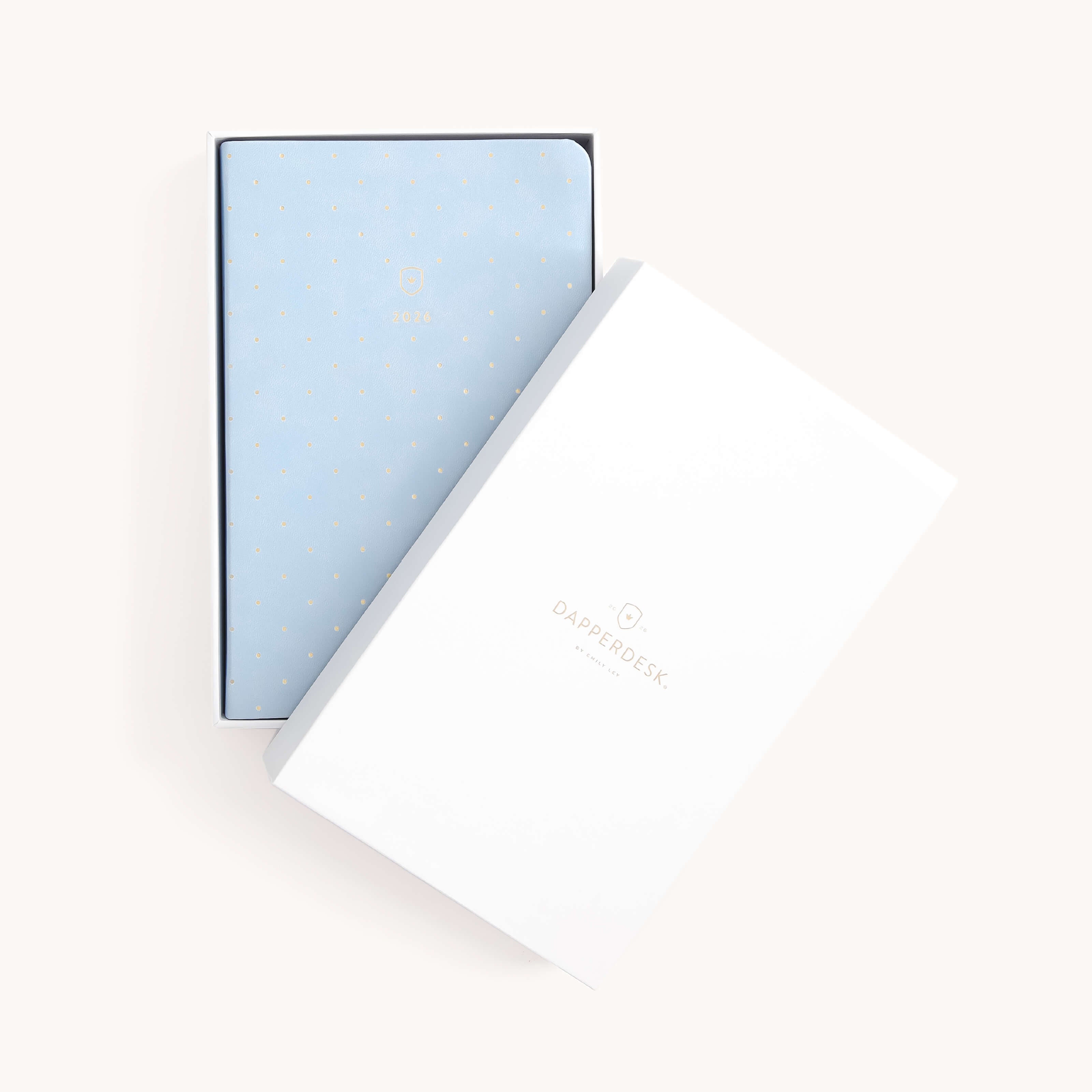 The 2026 Bookbound Daily Dapperdesk Planner - Carolina Dot by Simplified, a light blue 12-month planner with a subtle dot pattern and leatherette cover, rests in an open white Dapperdesk box. A gold emblem and text adorn the cover.