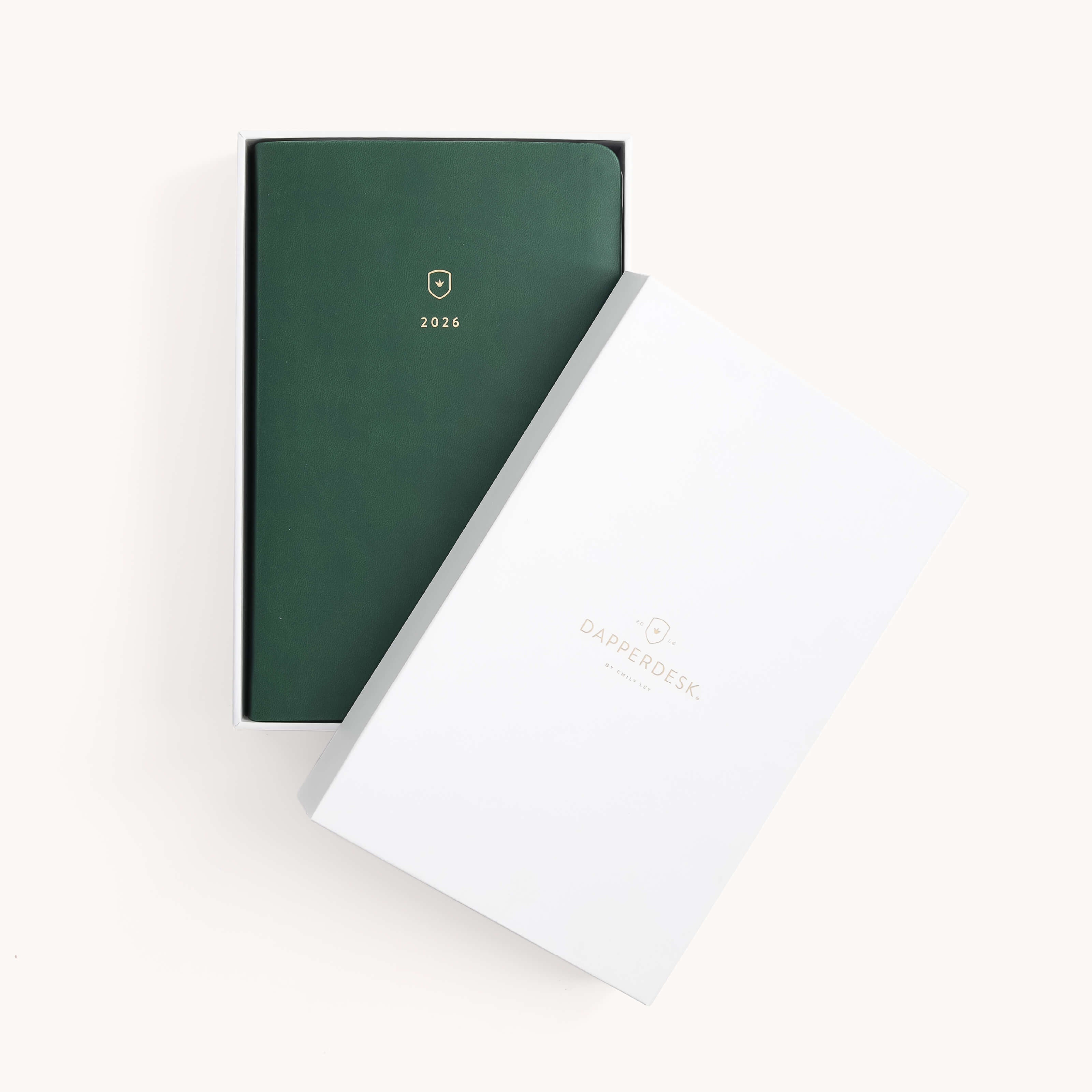 The 2026 Bookbound Daily Dapperdesk Planner - Evergreen by Simplified, featuring a dark green leatherette cover with gold details, rests in a white box with DAPPERDESK and a gold emblem on the lid, matching the planners own design.