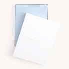 The 2026 Bookbound Weekly Dapperdesk Planner - Carolina Dot by Simplified, featuring a light blue leatherette cover with gold dots, is partially covered by a white folder with Dapperdesk in gold on the front.