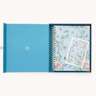 An open blue box holds a Simplified 2026 Spiral Weekly Simplified Planner - Faraway Land and a coordinating notecard, both adorned with floral and butterfly designs on a light blue background.