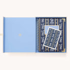 An open light blue box holds a navy blue 2026 Spiral Weekly Simplified Planner - Poppy Posies by Simplified, adorned with a white floral pattern and a matching card on top, both featuring elegant designs.