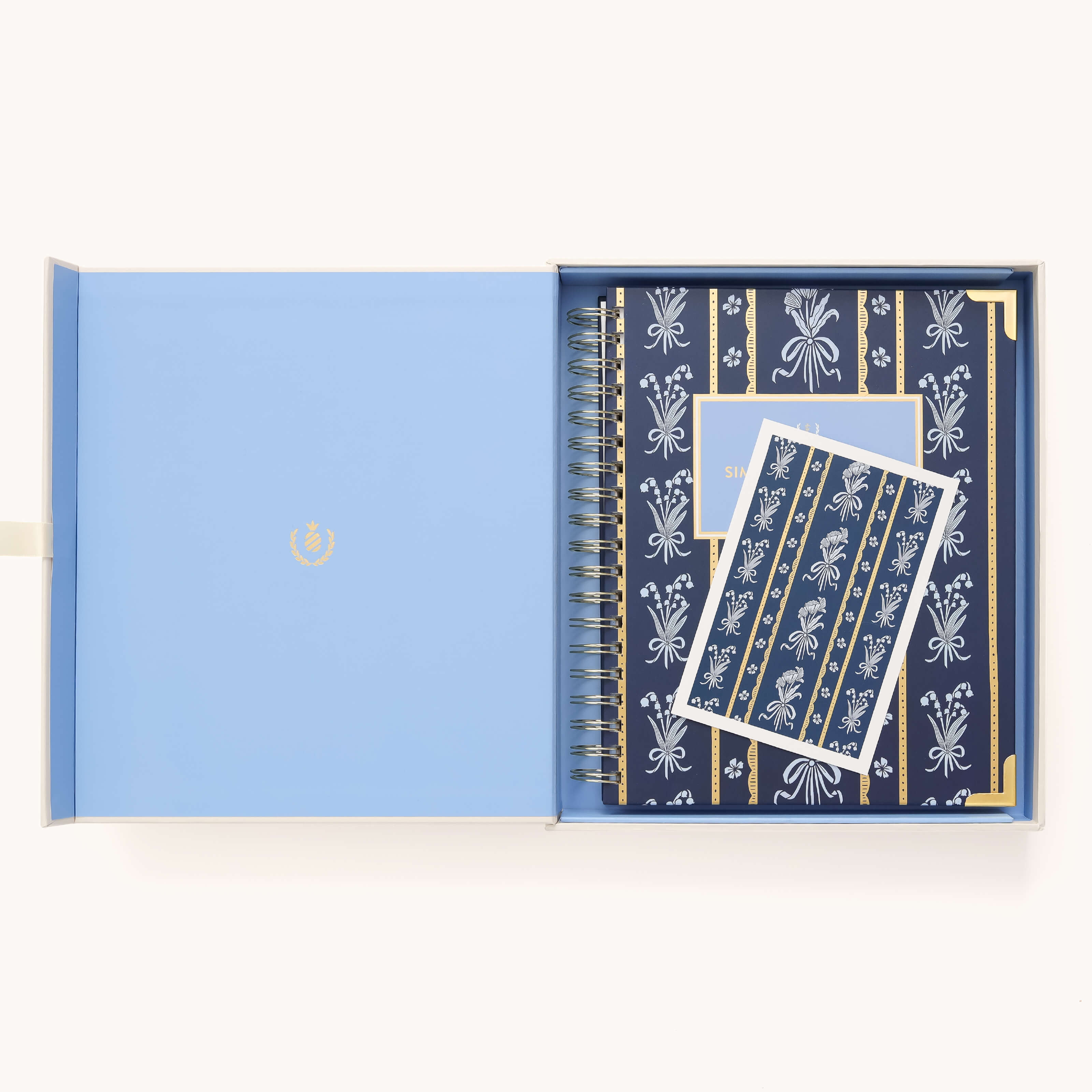 An open light blue box holds a navy blue 2026 Spiral Weekly Simplified Planner - Poppy Posies by Simplified, adorned with a white floral pattern and a matching card on top, both featuring elegant designs.