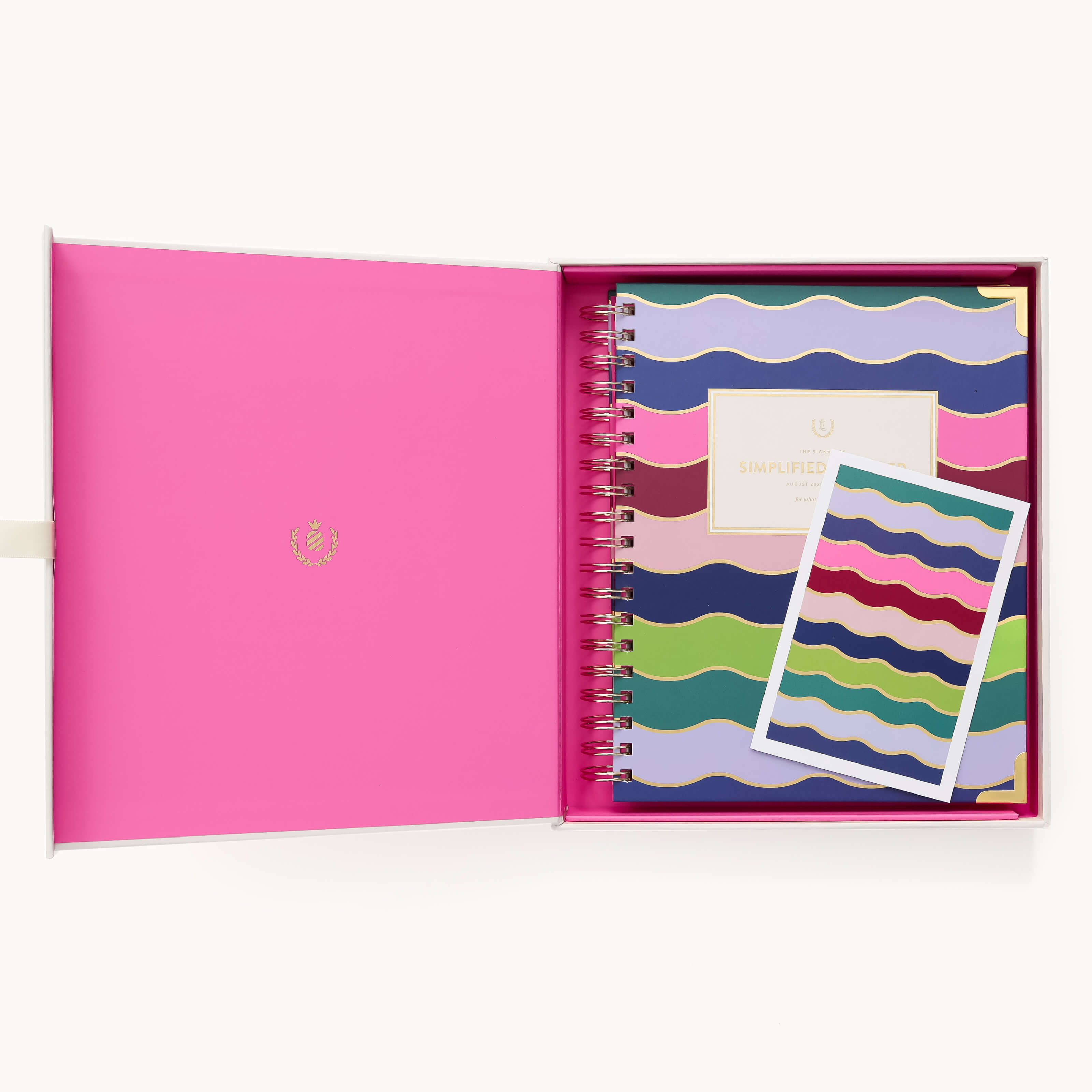 An open pink box displays the 2026 Spiral Weekly Simplified Planner – Rainbow Row by Simplified, with wavy blue, green, pink, and purple stripes, alongside a matching notecard in the same vibrant design.