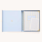 An open light blue box holds the 2026 Spiral Weekly Simplified Planner - Serene Stripe by Simplified, featuring blue and white stripes with gold accents, plus a coordinating notecard, both neatly arranged inside.