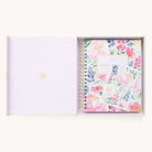 An open box displays the Simplified 2025-2026 Spiral Weekly Simplified Planner in Cottage Floral, showcasing pink, red, and purple flowers with elegant gold foil details. Inside, a matching bookmark and card lie on a pristine white background, completing the charming ensemble.